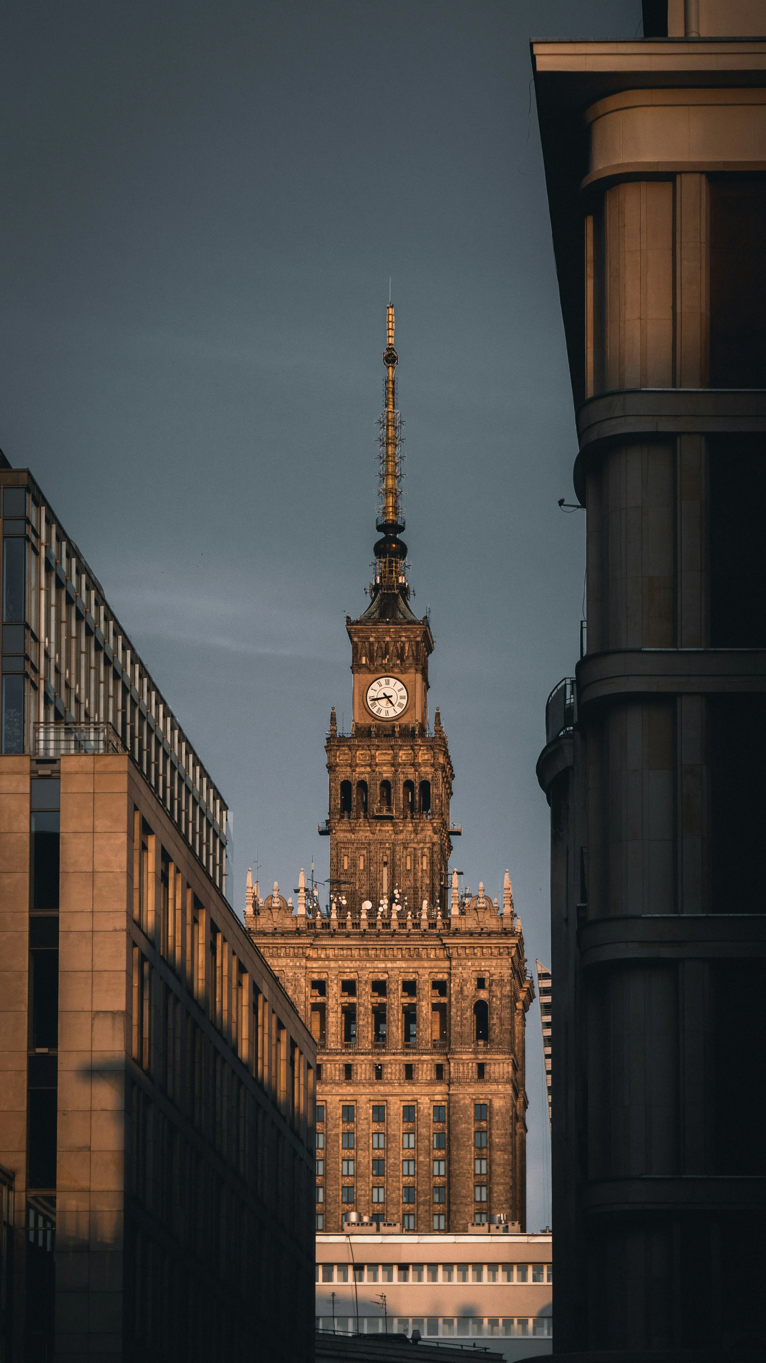 Tall historic clock tower of the Palace of Culture and Science in Warsaw, Poland, with buildings on either side and a clear sky background.