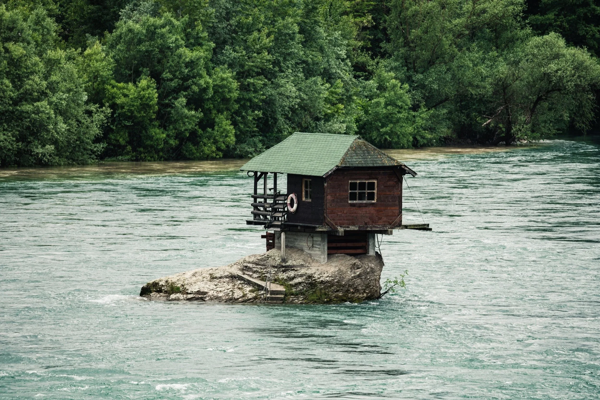 Small house built on a rock in the middle of a river, surrounded by green trees.