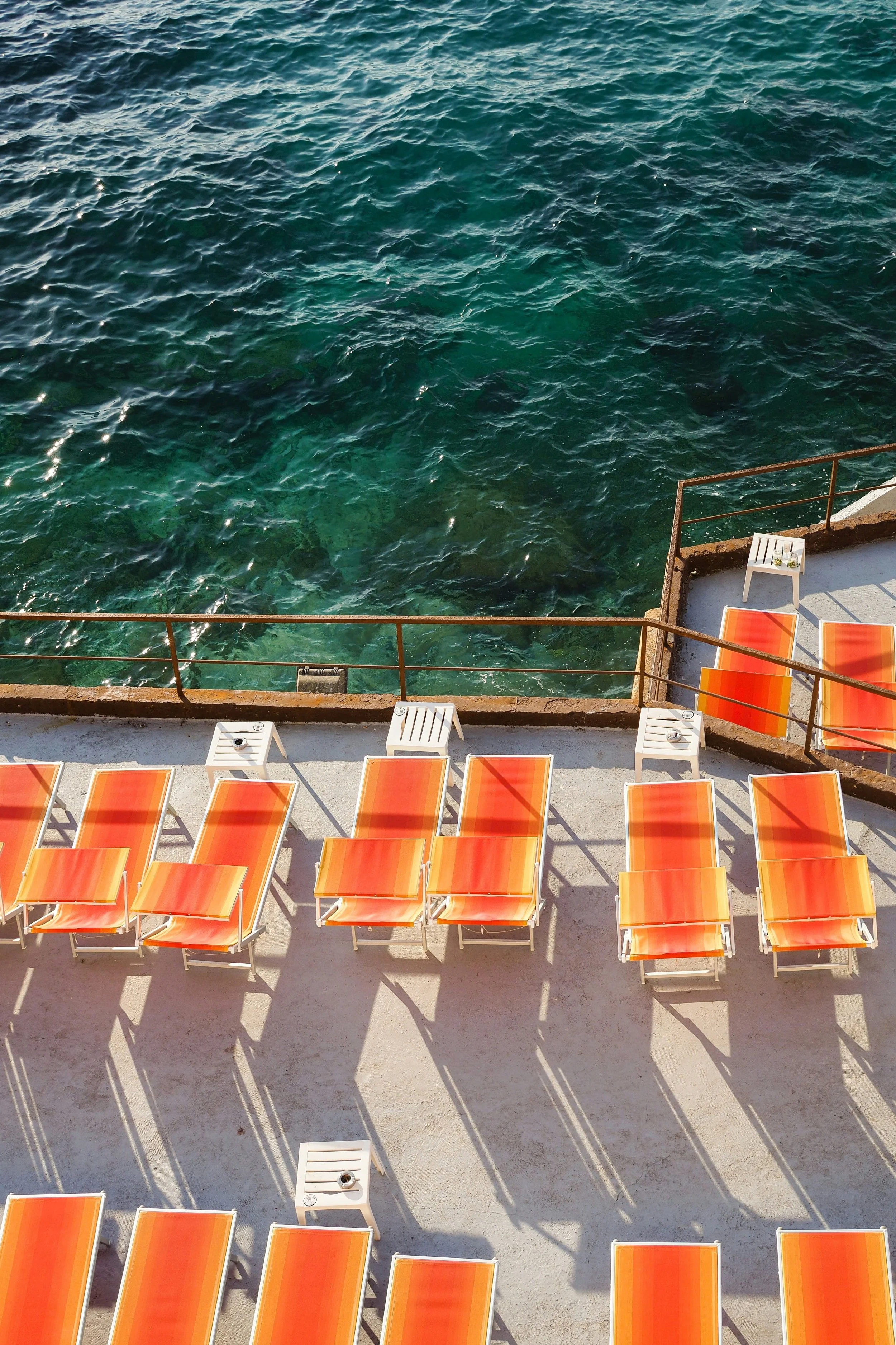 Empty orange lounge chairs and white tables on a balcony overlooking clear blue-green water.