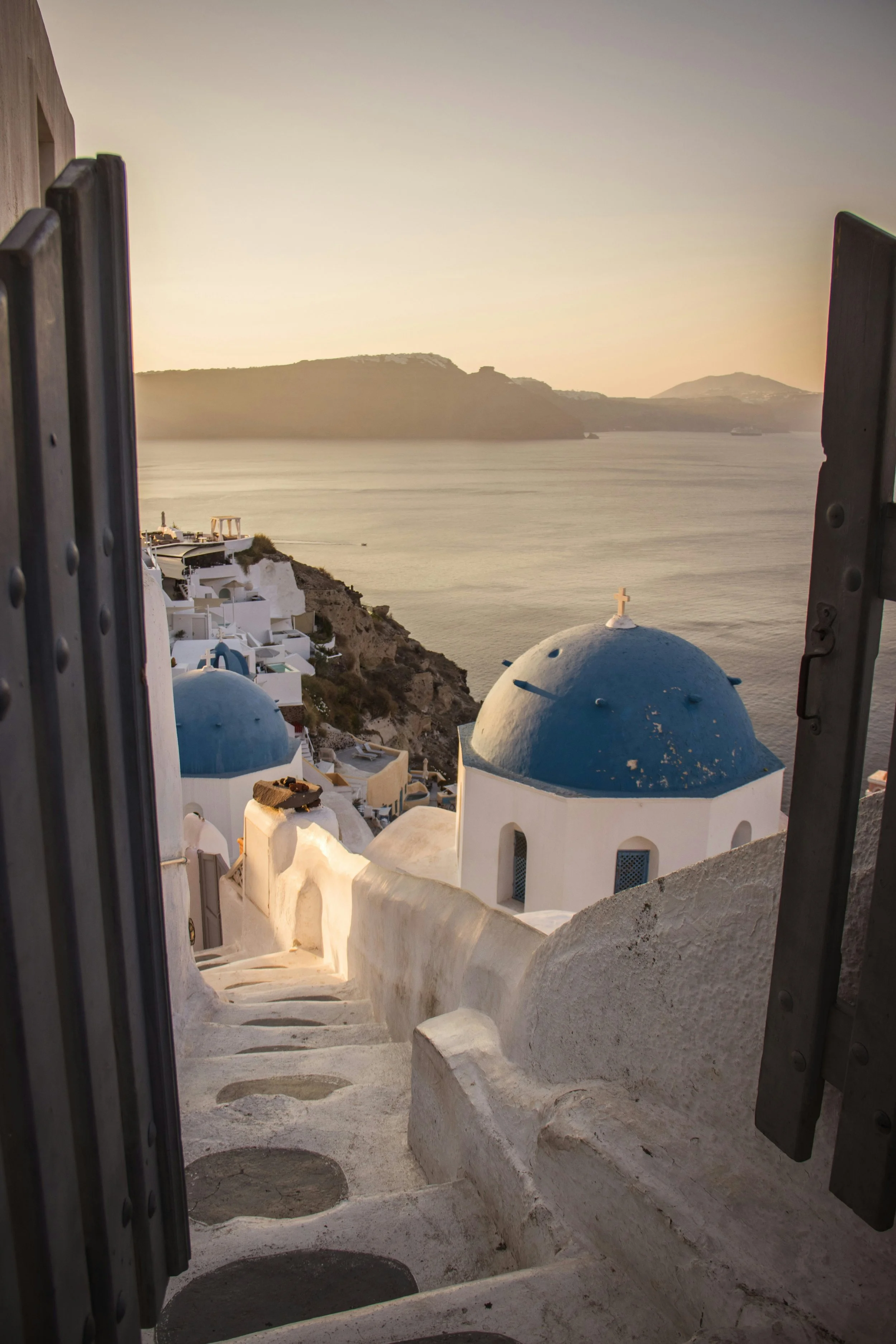 White stairs leading to a view of white buildings with blue domes on a hillside overlooking the water, with hills in the background at sunset.