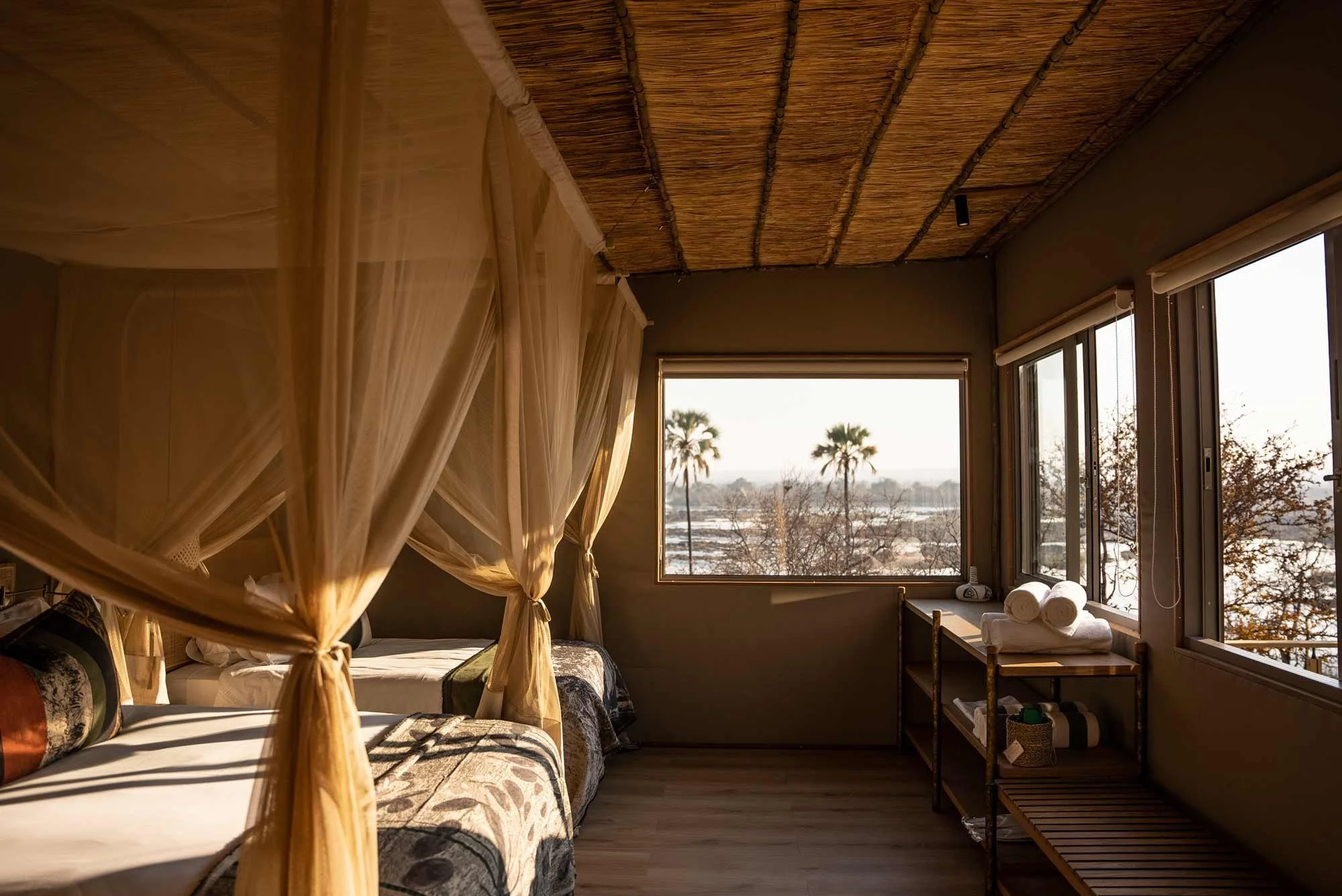 A cozy bedroom with a four-poster bed with sheer curtains, wooden flooring, and large windows overlooking palm trees and a scenic landscape.