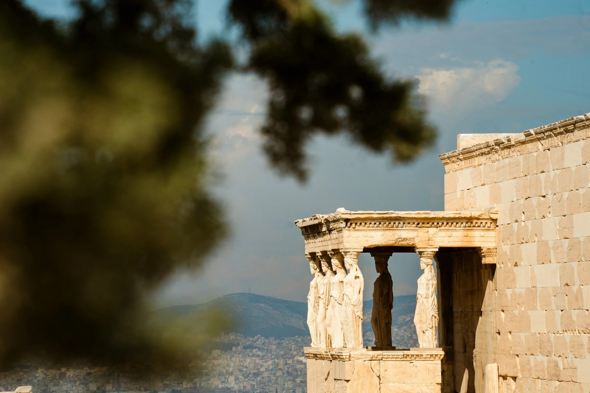 Ancient Greek temple ruins with carved columns, with a blurred tree in the foreground and a cityscape with hills and a cloudy sky in the background.