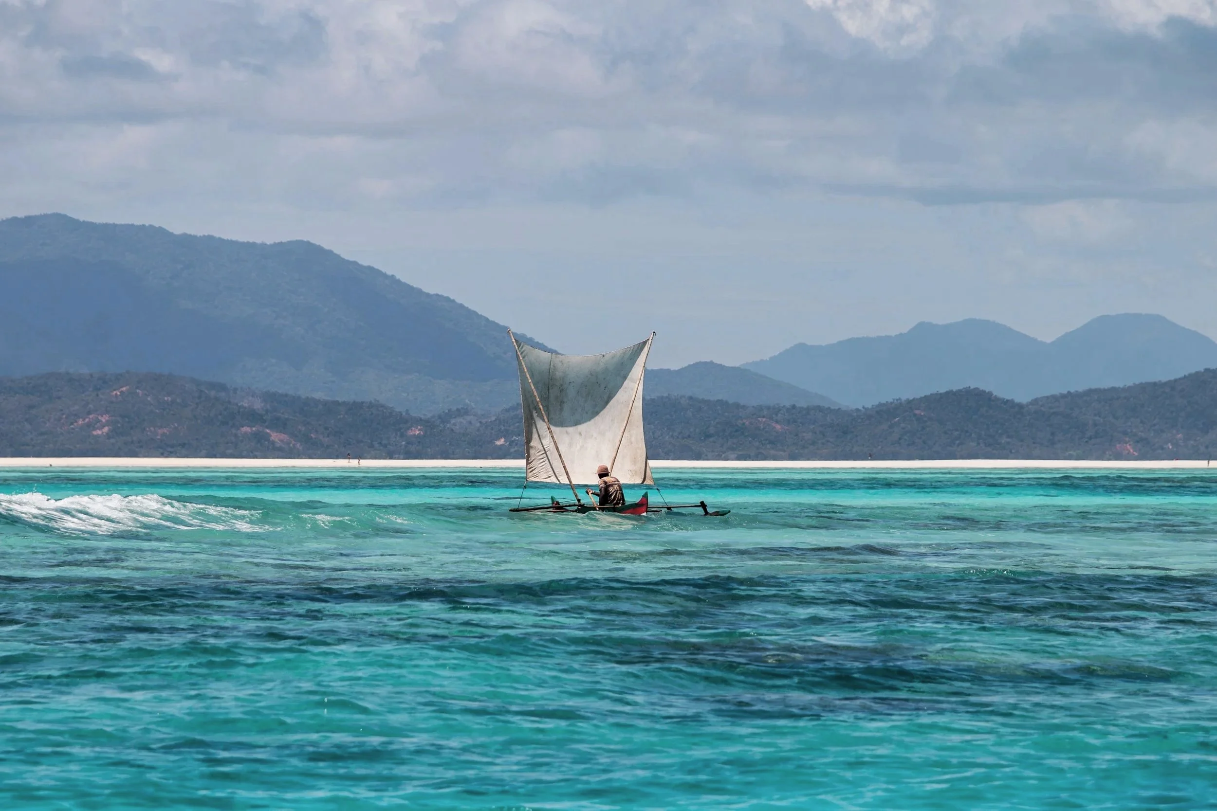 A small sailboat with a single sail is on turquoise water, with mountains and a cloudy sky in the background.