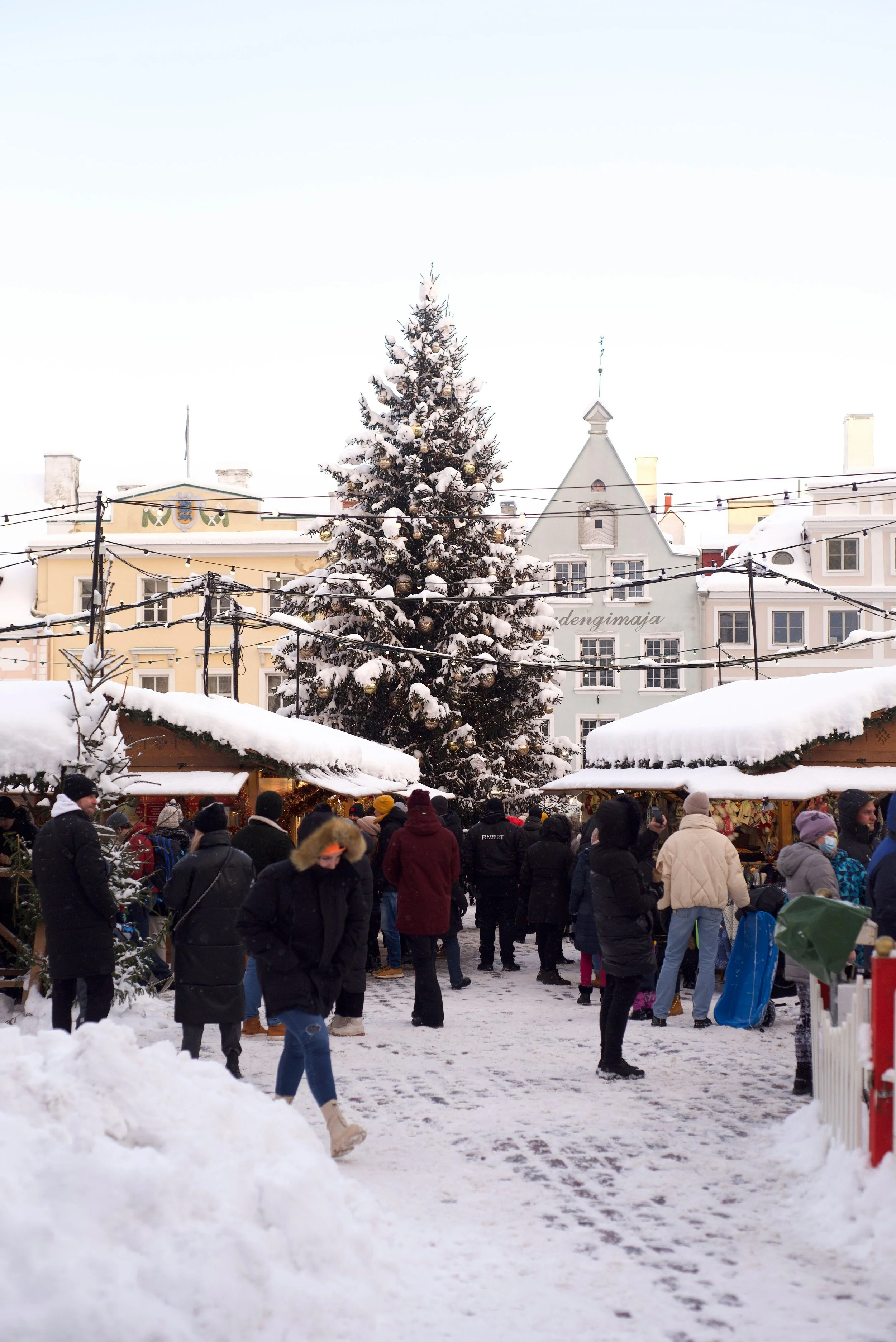 Snow-covered Christmas market with a decorated Christmas tree, wooden stalls, and people bundled up in winter clothing.