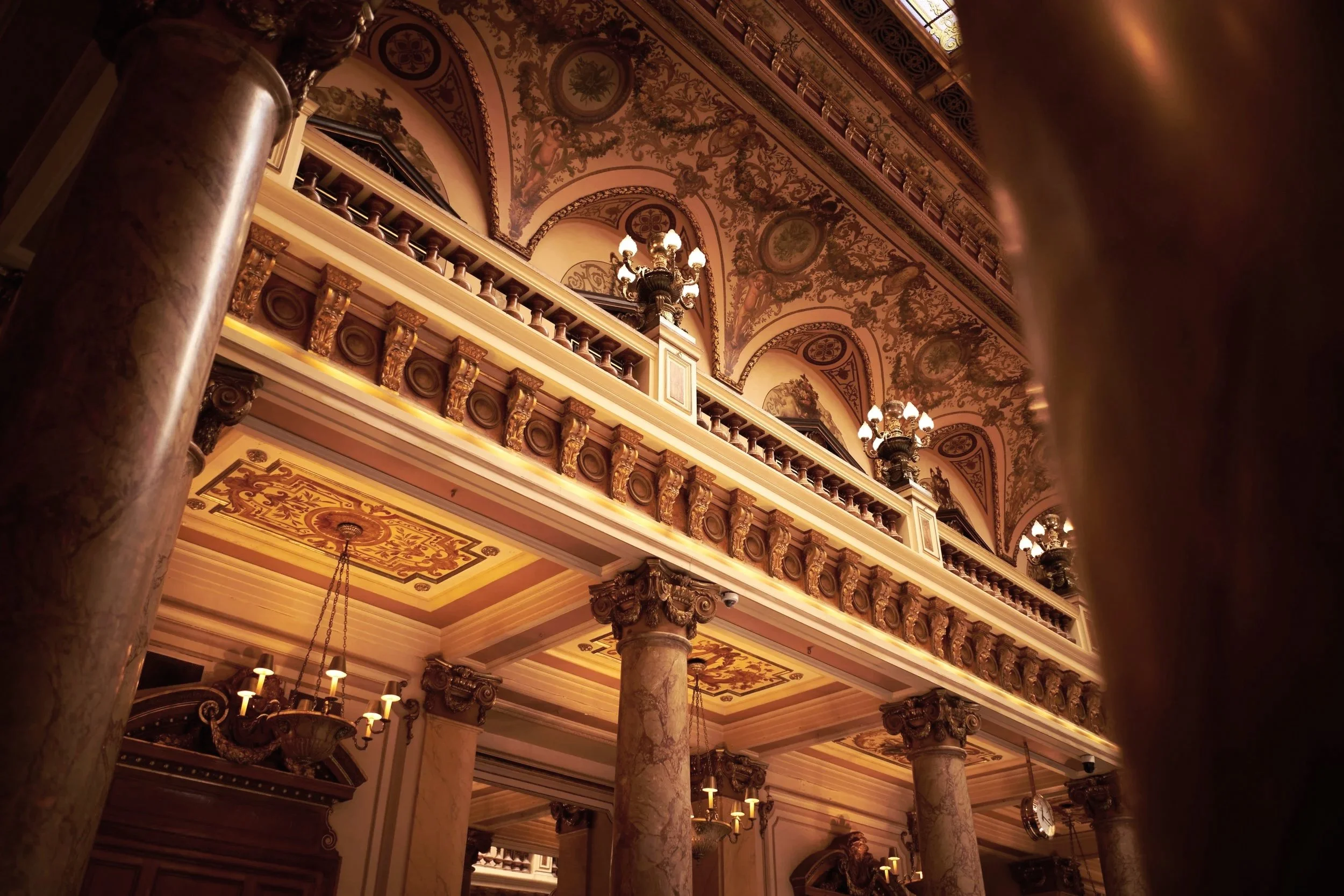 Elegant interior of a historic building featuring ornate columns, intricate ceiling details, chandeliers, and decorative lighting fixtures.