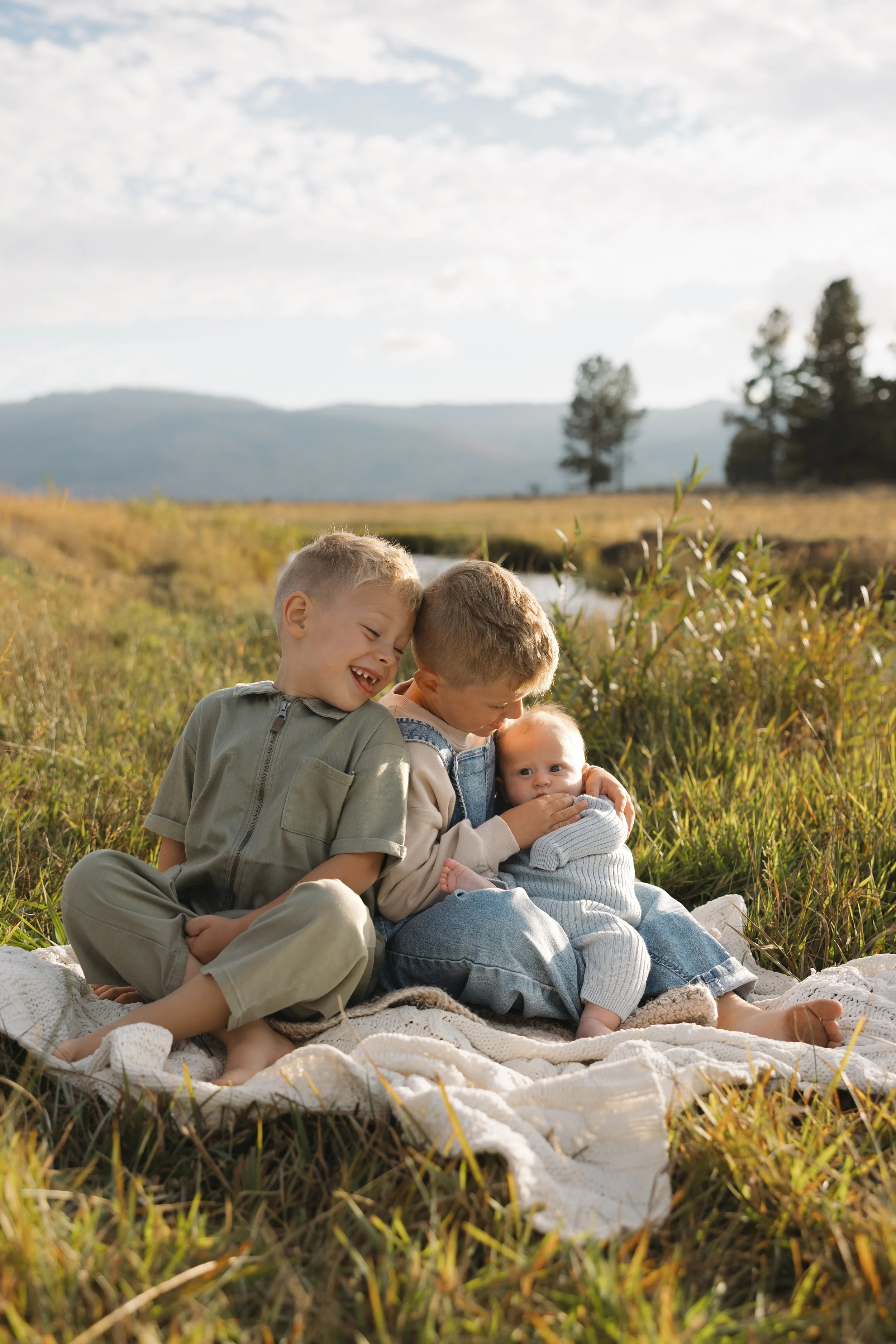 Four children sitting on a blanket in a grassy outdoor field with mountains and trees in the background, enjoying each other's company on a sunny day.