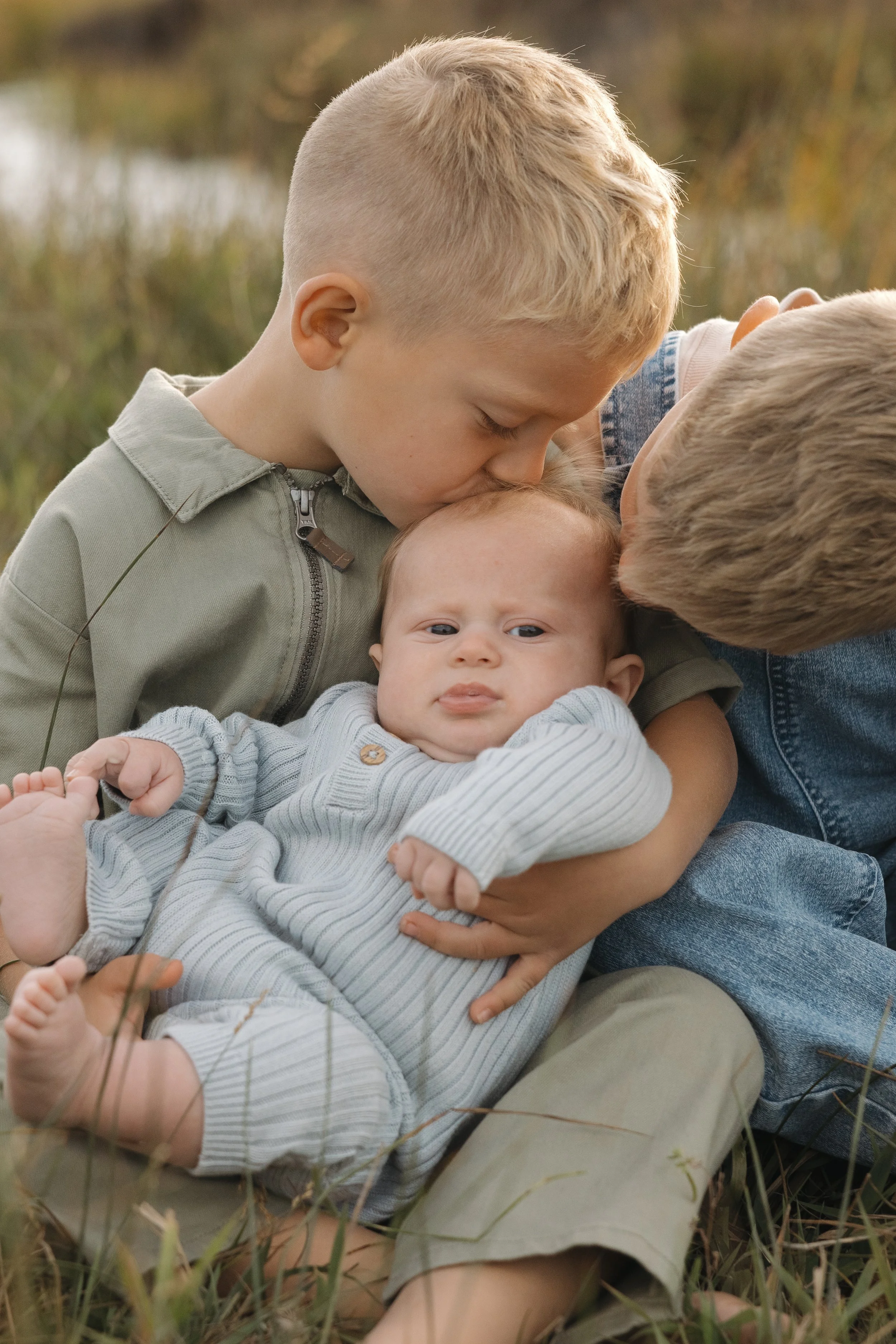 Two children holding a baby outside in a grassy area during daytime, with one child kissing the baby's head.