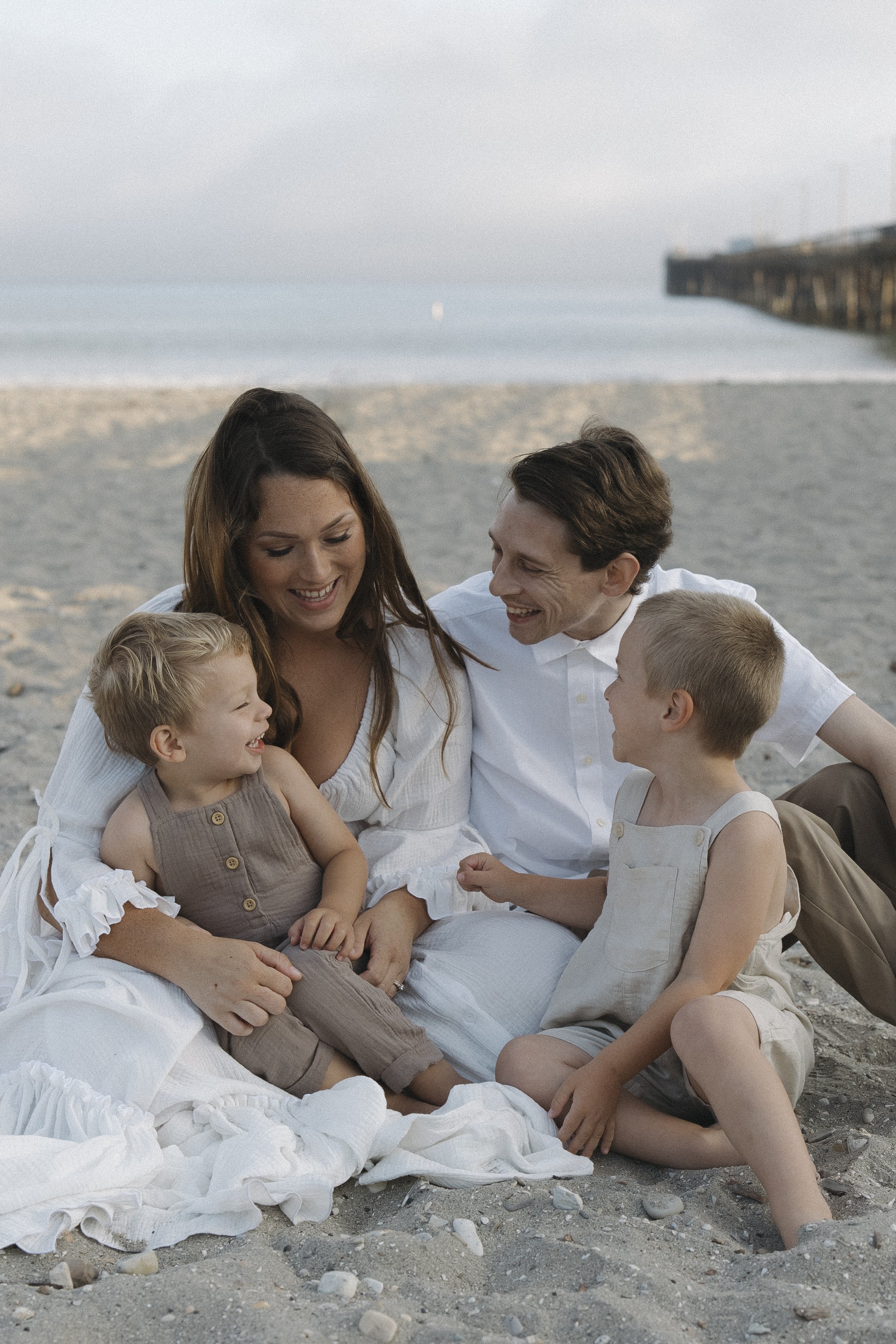 A family of five sitting on the beach, smiling and enjoying each other's company during sunset, with the ocean and a pier in the background.