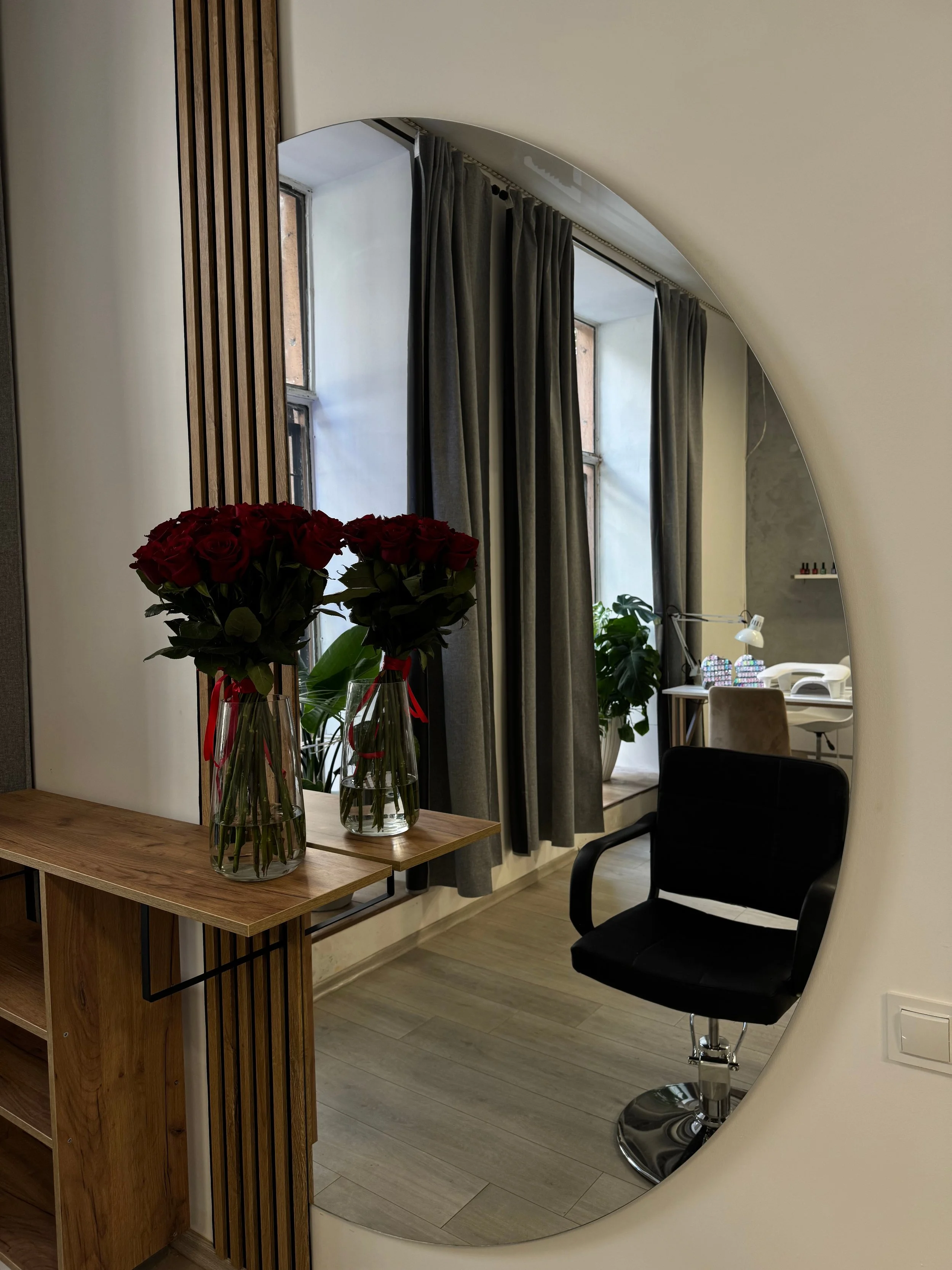 An interior space with a mirror reflecting a room containing a black salon chair, a desk, a lamp, and some decorations, with two vases of red roses on a wooden table in the foreground.