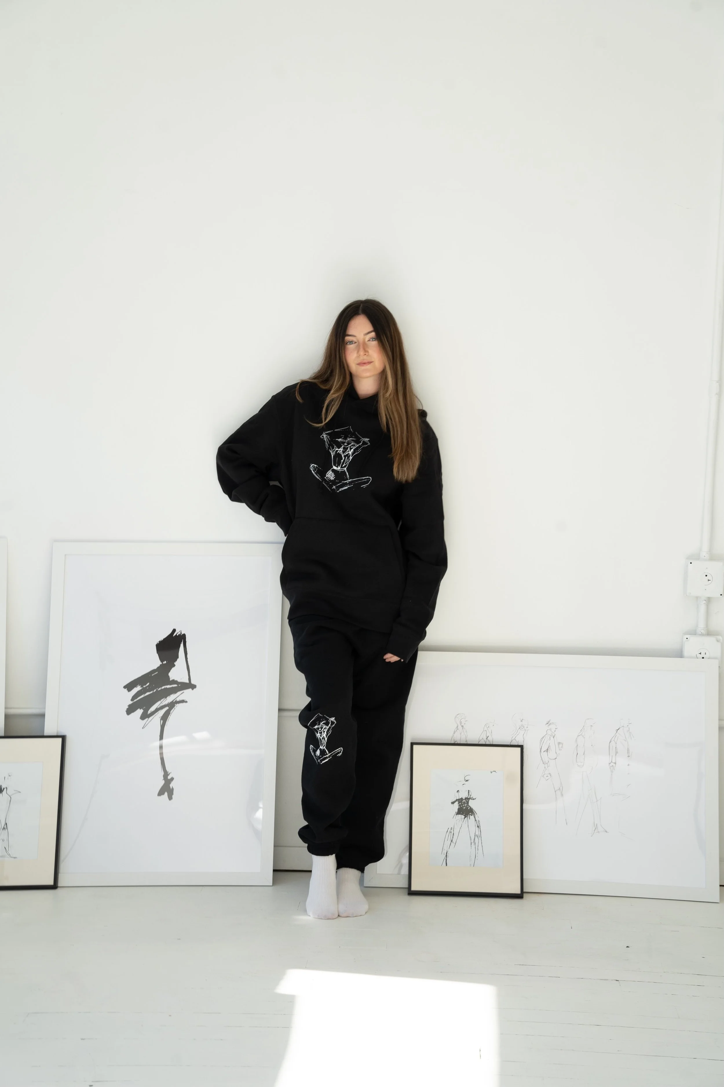 A woman in a black hoodie and sweatpants with white socks stands against a white wall, surrounded by framed artwork and sketches on the floor and leaning against the wall.