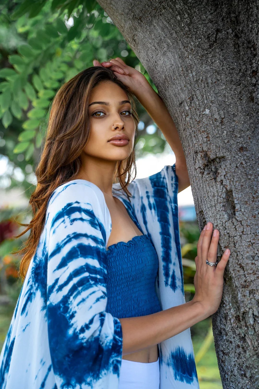 A woman with shoulder-length black hair standing outdoors in a tropical garden with large green leaves and purple flowers, wearing a sheer maroon crochet top and wide-leg tie-dye pants in white and purple, and beige high heels.