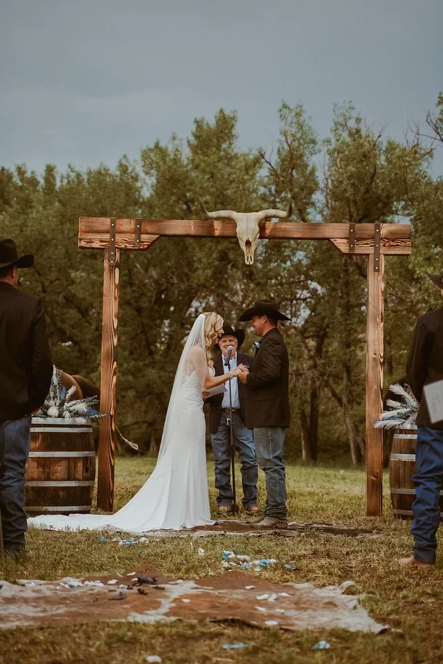 A couple getting married under a rustic wooden arch with a cow skull decoration, surrounded by friends, outdoors with green trees in the background, during daytime.