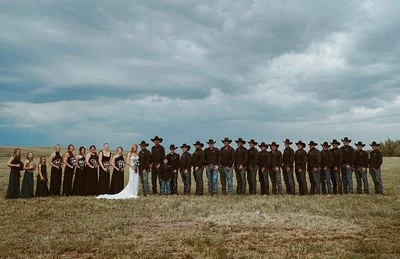 A large group of people dressed in period costumes, with women in dresses and men in cowboy hats, standing in an open field under a cloudy sky, possibly a reenactment or historical event.