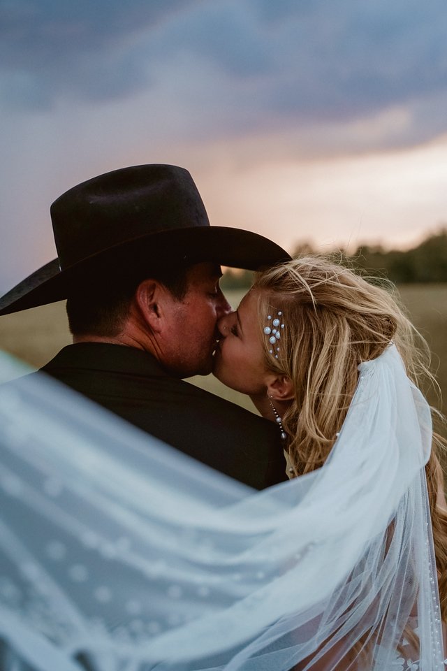A couple, a man in a black cowboy hat and a woman with long blonde hair and pearl accessories, sharing a kiss outdoors during sunset.