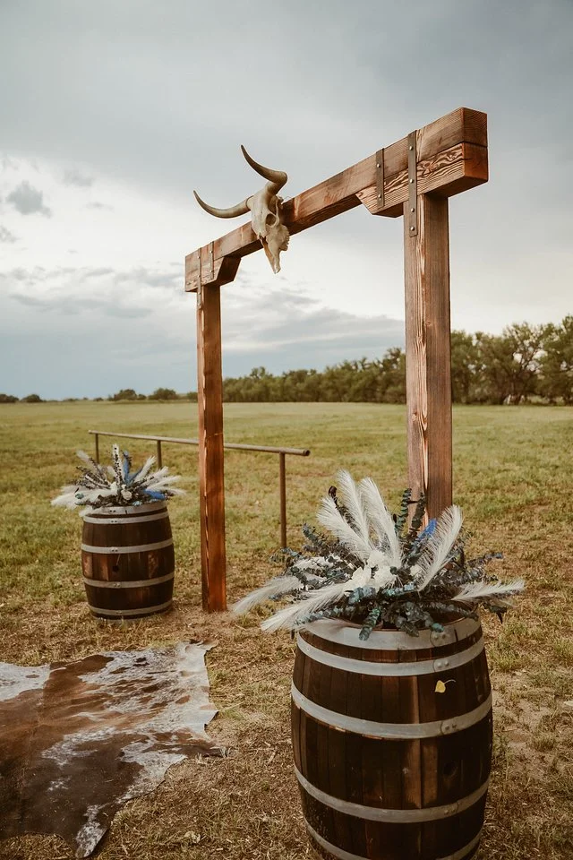 Outdoor rustic setup with a wooden arch, decorated with a skull of a longhorn steer mounted on top, complemented by barrels filled with white and blue decorative feathers and foliage; cloudy sky overhead.
