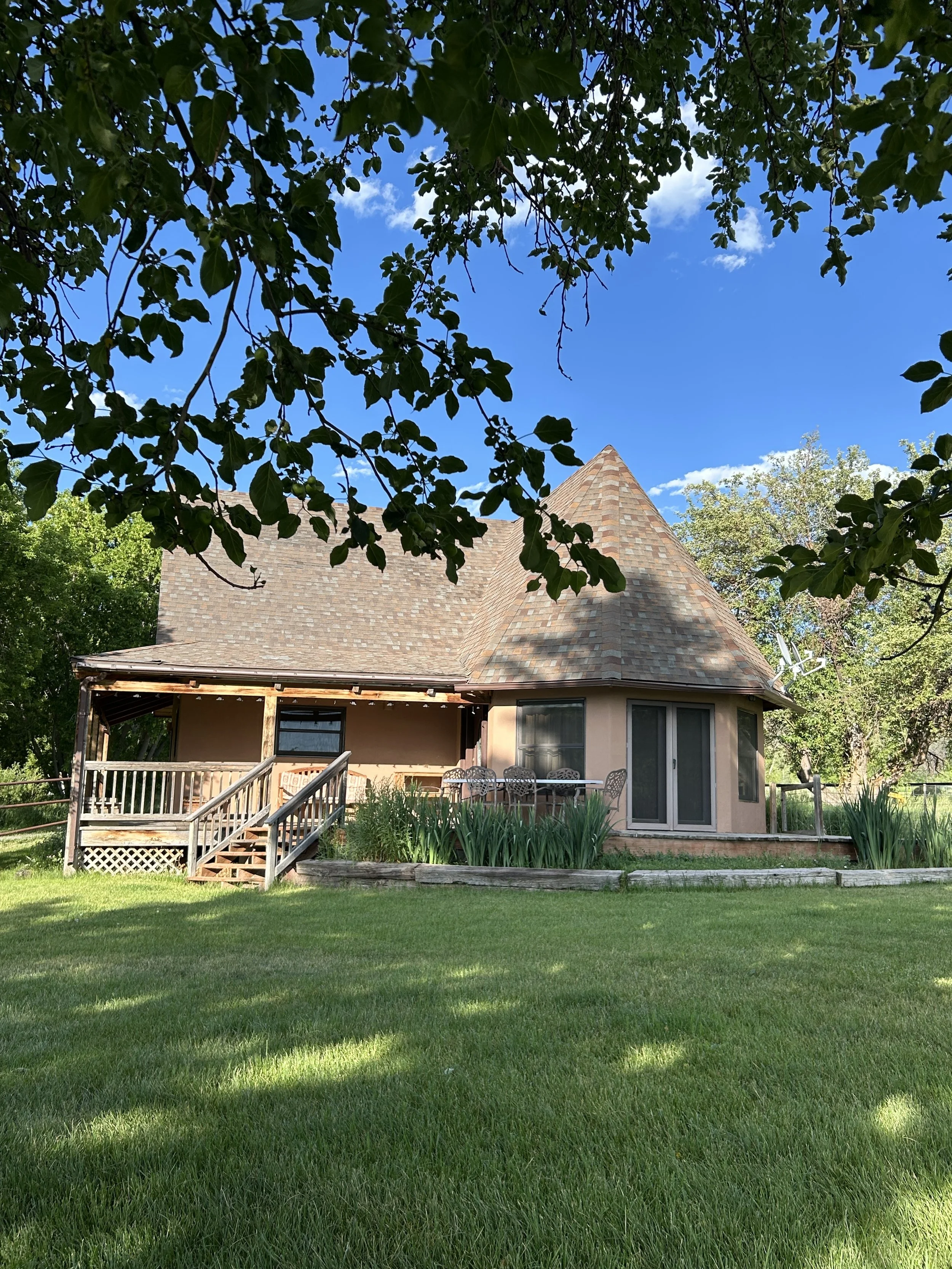 A house with a round turret and a wooden front porch, surrounded by a green lawn and trees under a blue sky with some clouds.