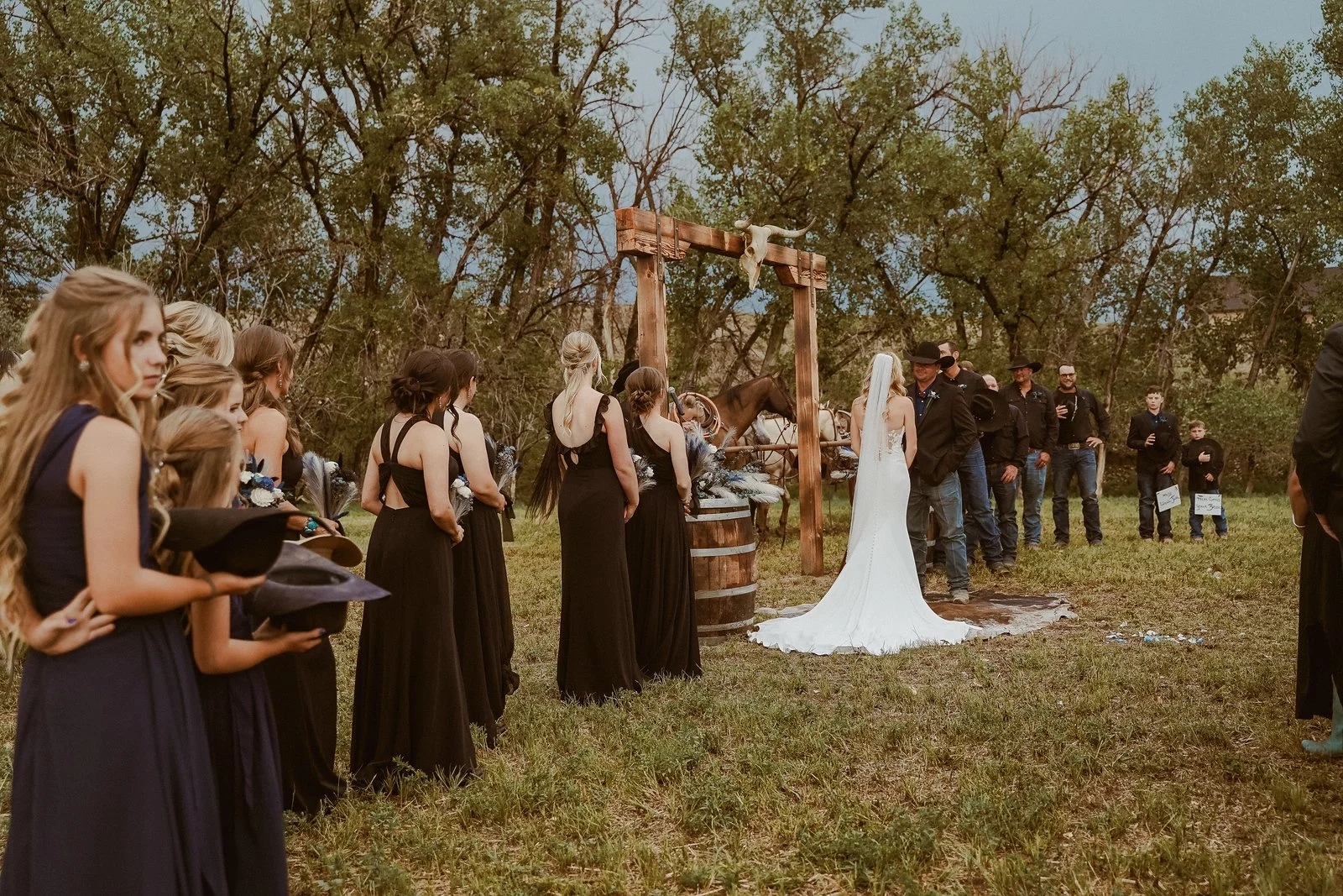 A outdoor wedding ceremony with bridesmaids in black dresses and groomsmen in black attire, a bride in a white wedding gown, and a groom standing under a wooden arch with a white cloth train on the ground, surrounded by trees.