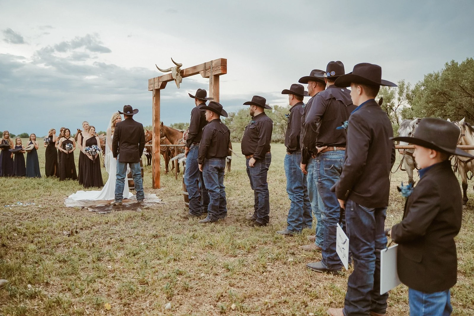 A wedding ceremony outdoors with a bride and groom standing in front of an officiant, surrounded by bridesmaids and groomsmen dressed in black jackets and cowboy hats. The groomsmen are lined up, standing next to horses. The setting is a grassy field with trees and cloudy skies in the background.