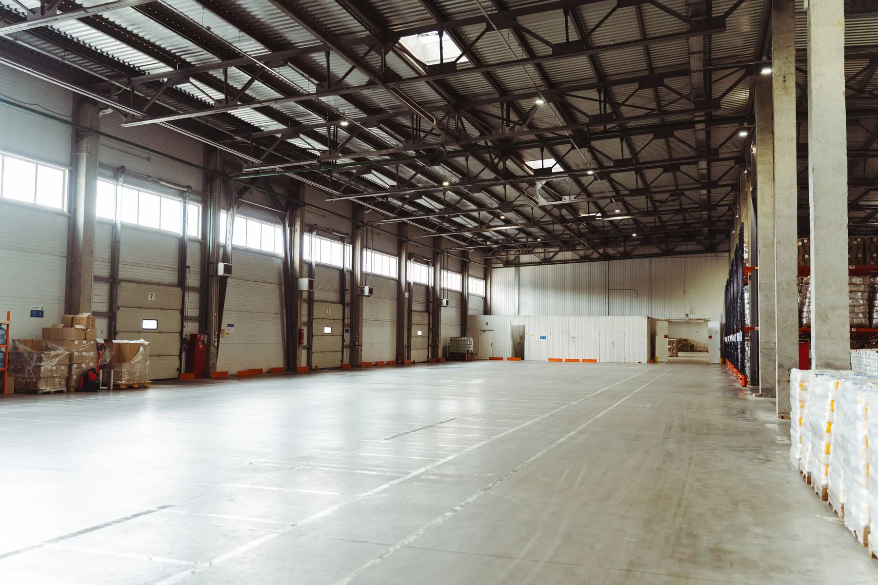 Empty warehouse with large garage doors, concrete floor, storage shelves, and pallets on the right side.