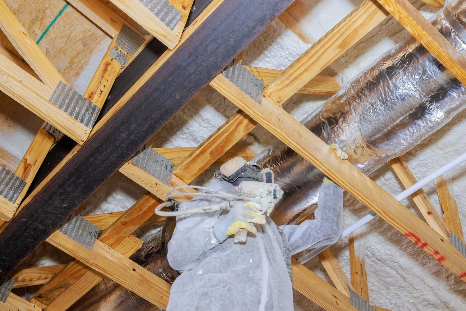 View of a worker's legs and hand holding a nail gun, working on wooden framing in an attic or ceiling space, with insulation and ductwork visible.