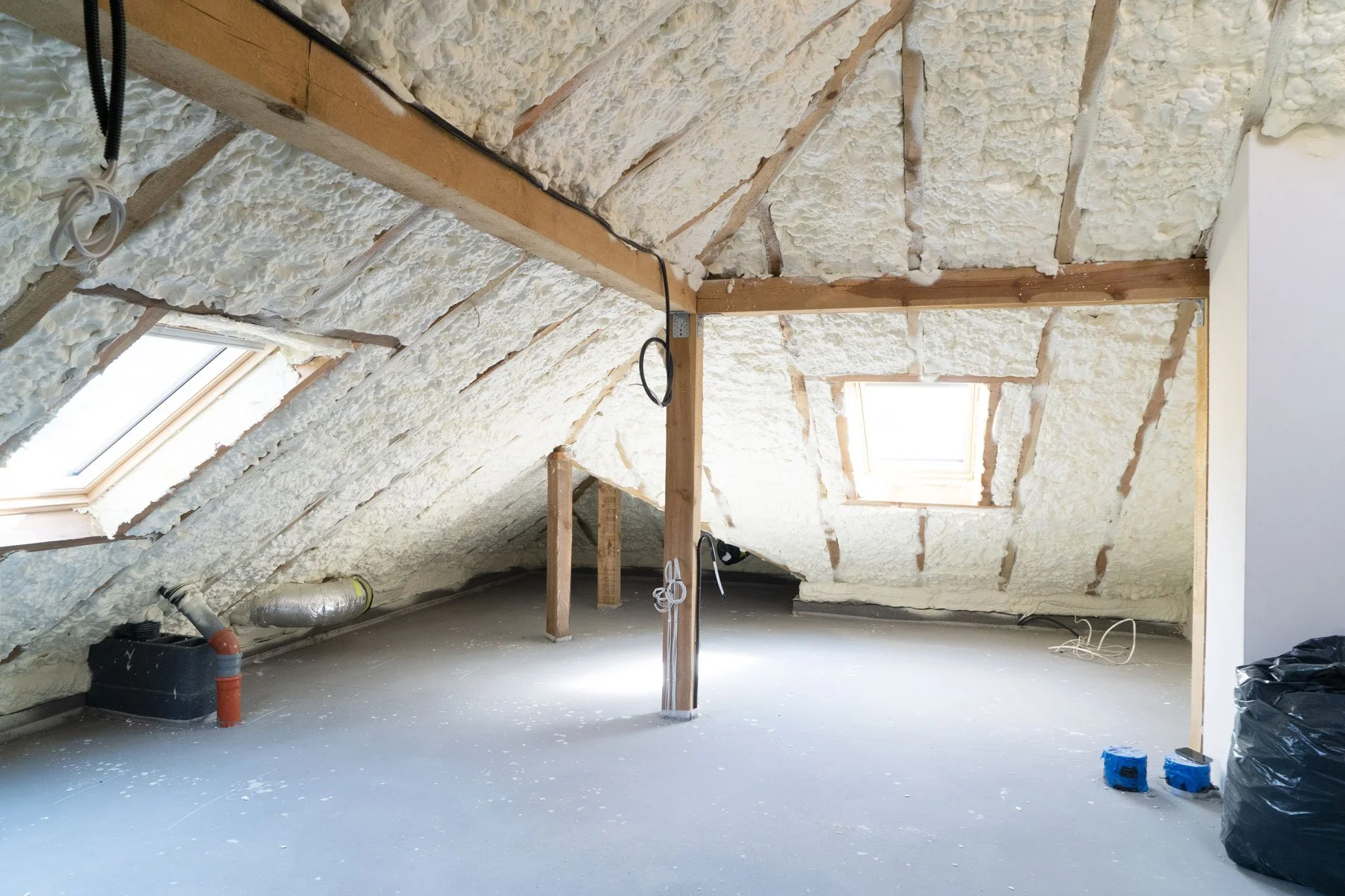 An attic room under construction with insulation spray foam on the sloped ceiling. There are two skylight windows, exposed wooden beams, and some visible pipes. The room has a gray concrete floor with some miscellaneous items like a black bag and small blue objects.