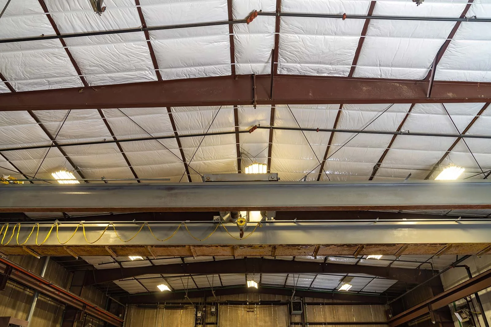 Interior view of an industrial warehouse ceiling with metal beams, insulation, and lighting fixtures.