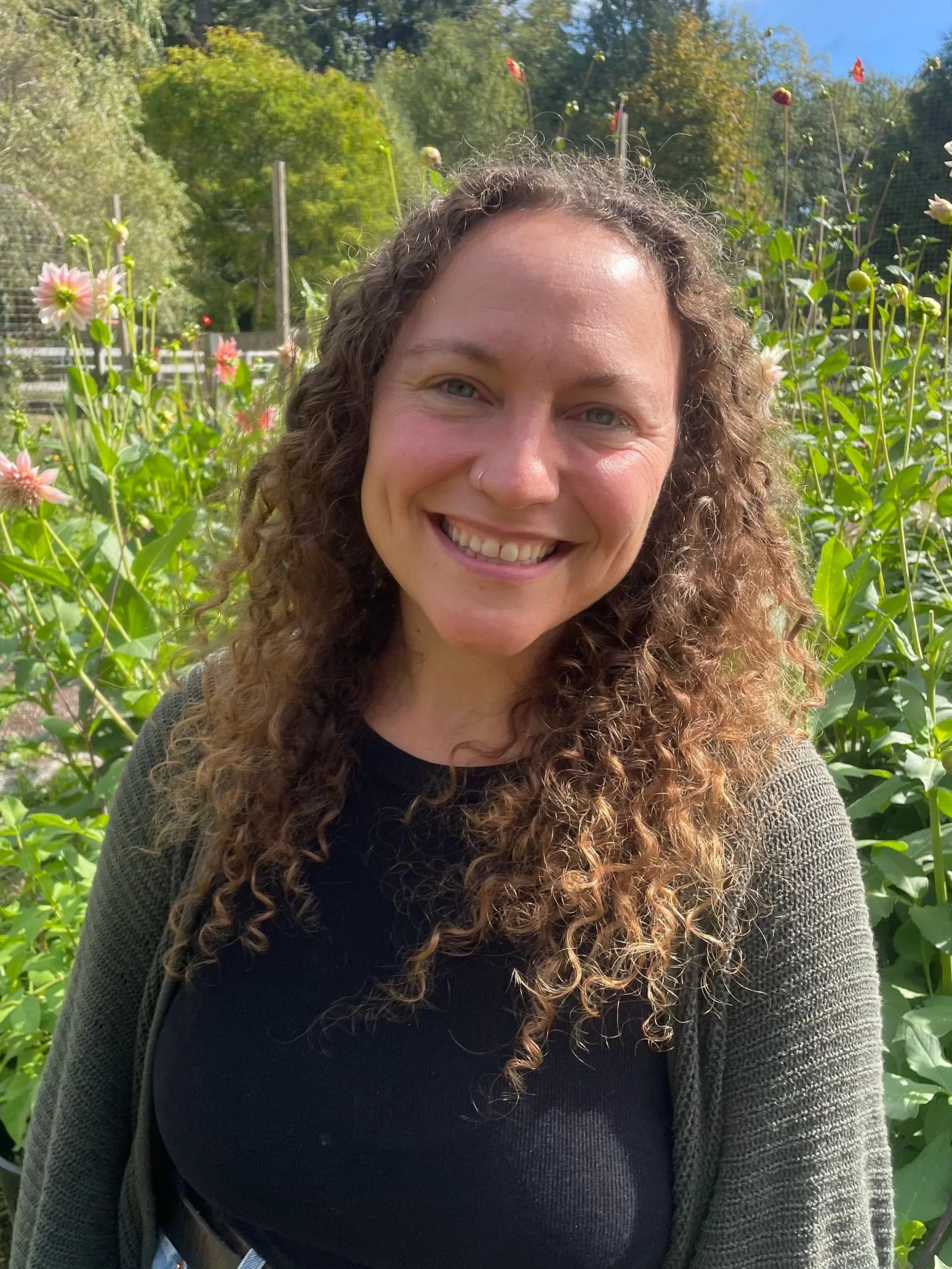 A smiling woman with curly brown hair and a nose ring standing in a garden surrounded by lush green plants and dahlia flowers rising taller than she is.