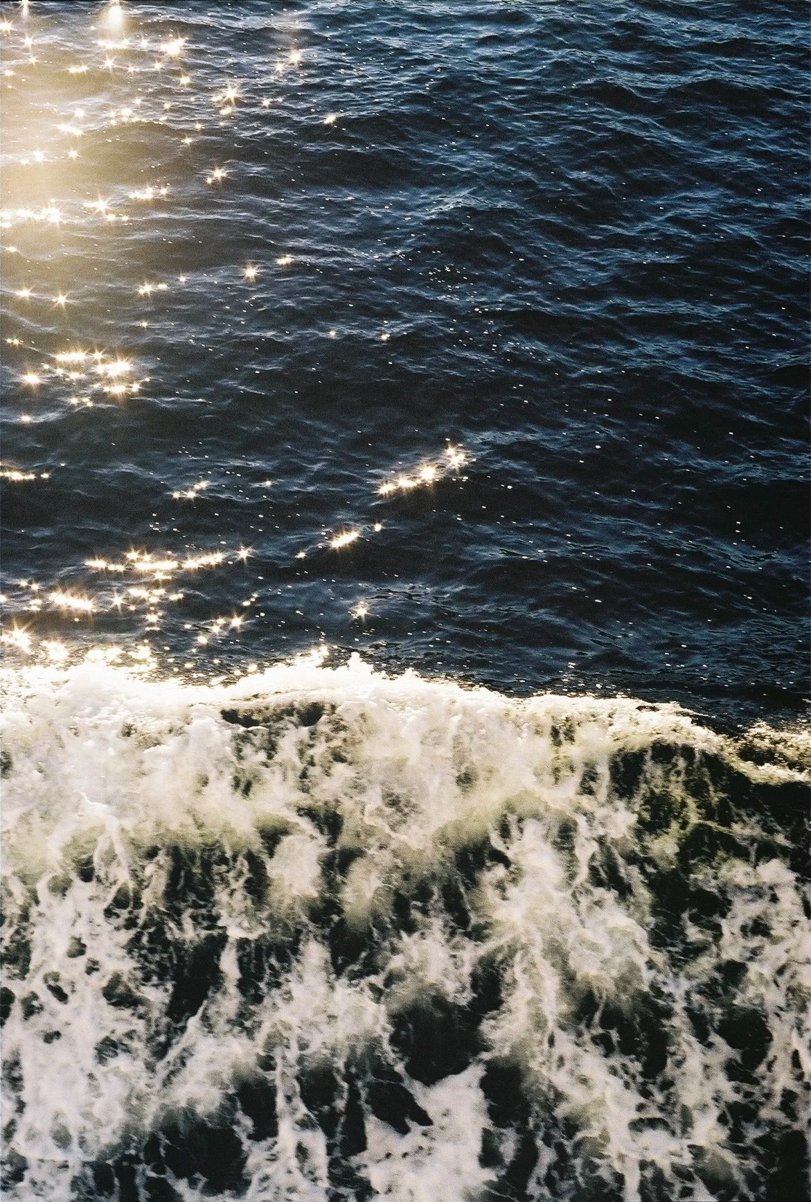 Close-up photo of dark blue ocean water with sunlight reflecting off the surface and white foamy wake at the bottom of the image.