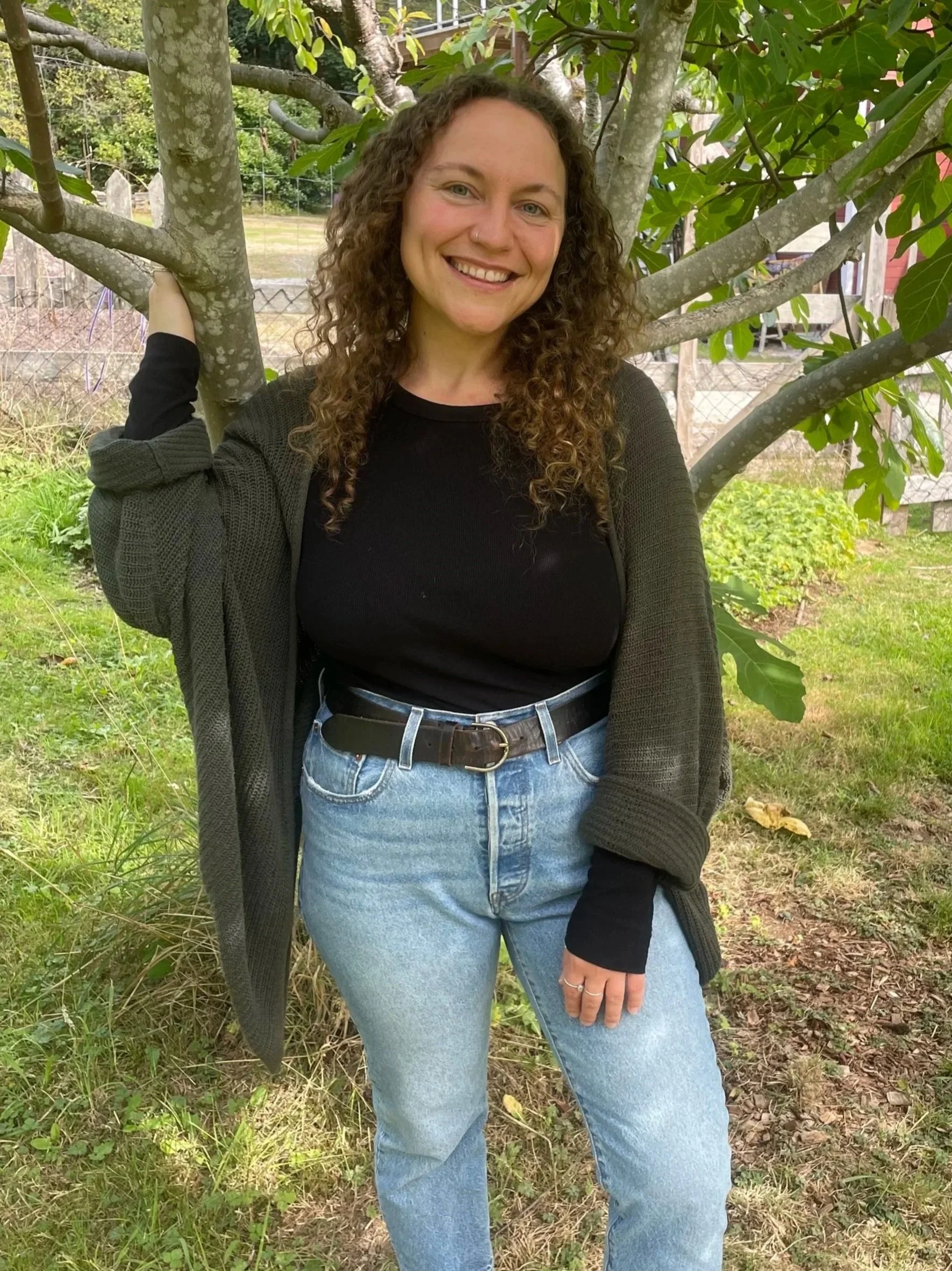 A woman with curly brown hair smiling outdoors, standing under a fig tree with large green leaves, wearing a black shirt, blue jeans, a green cardigan, and tan boots, in a yard with grass and a wooden fence.
