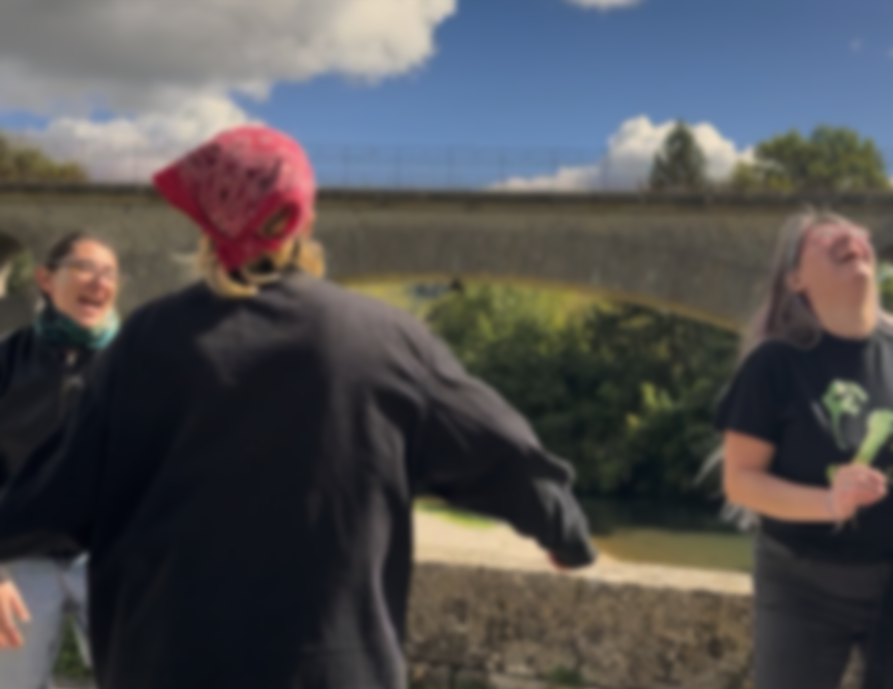 A non-binary people enjoying a cooking class outdoors, with two women in the center smiling and laughing while exchanging a friendly gesture, surrounded by cooking ingredients and utensils. queer joy, queer class, queer laughter.