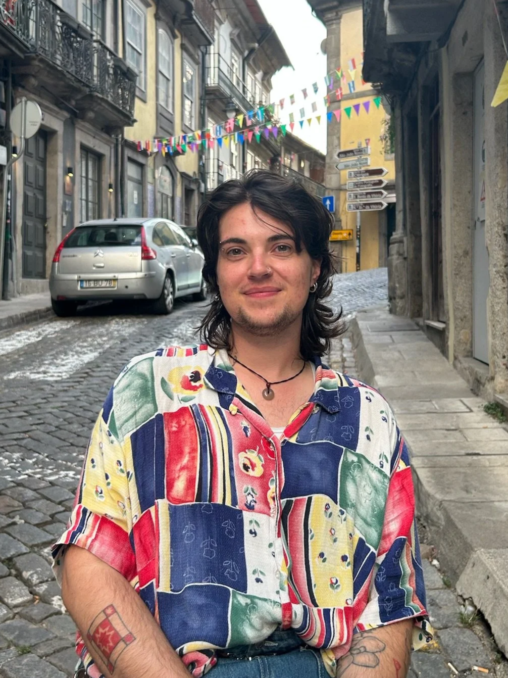 A person with dark, long hair, wearing a colorful patchwork shirt and earrings, standing on a cobblestone street in an old European city with colorful banners hanging overhead.