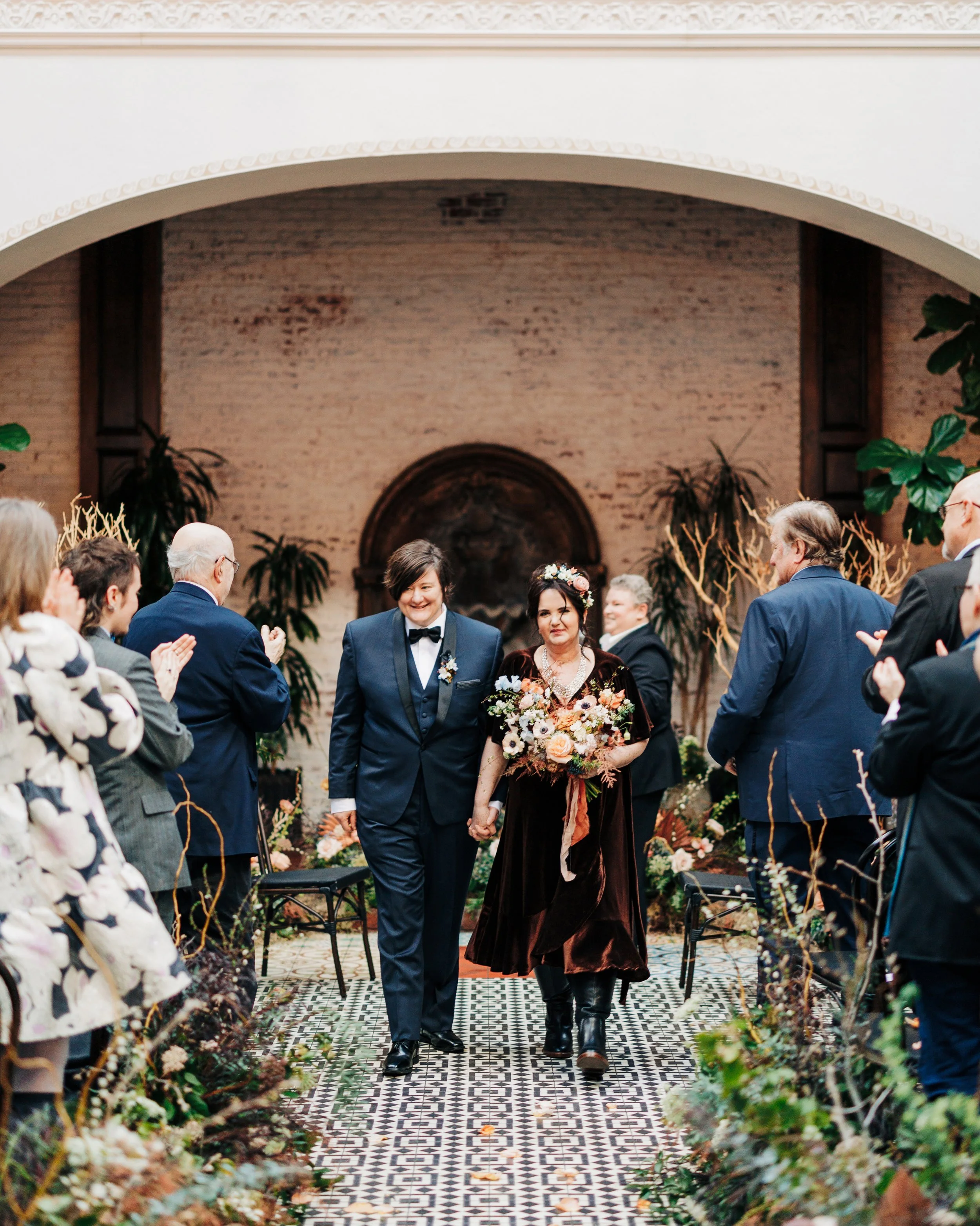 A couple of women are walking down the aisle of an indoor wedding ceremony, holding hands and smiling. The groom is wearing a dark blue tuxedo with a black bowtie, and the bride is dressed in a dark dress with floral decorations and a floral headband. Guests are standing on the sides, clapping and smiling. The setting is decorated with plants and flowers, with a brick wall and ornate ceiling details in the background.