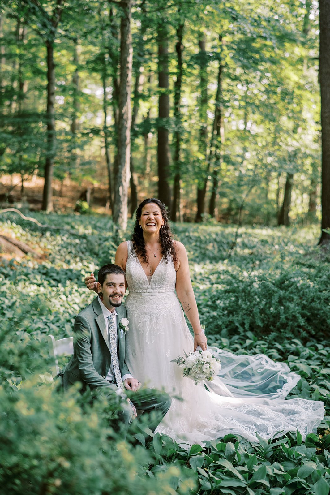 A bride and groom in wedding attire, sitting and standing in a lush green forest, smiling happily.