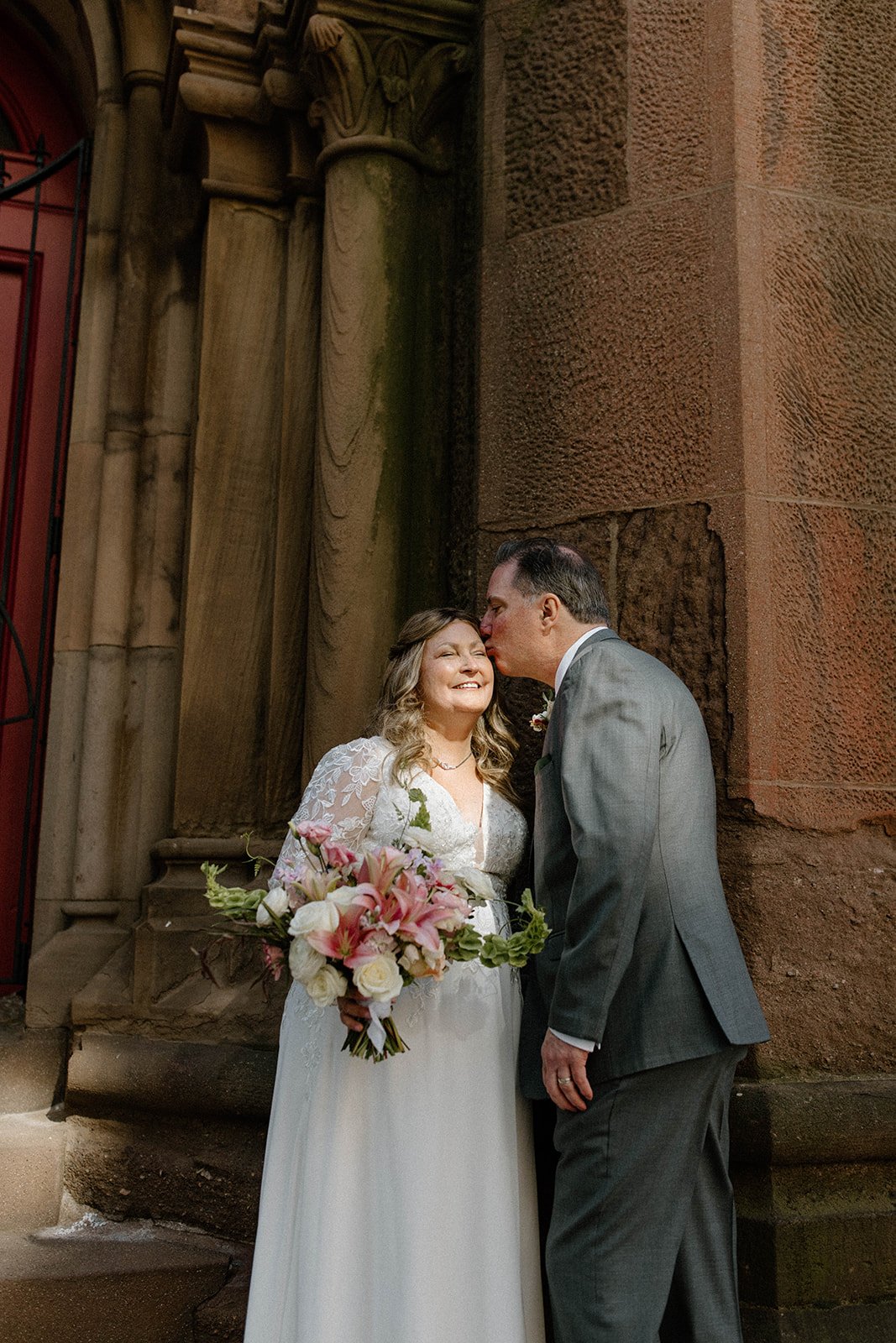 A bride and groom share a loving moment outside a historic stone building. The bride holds a bouquet of pink lilies and white roses, smiling as the groom kisses her on the cheek.