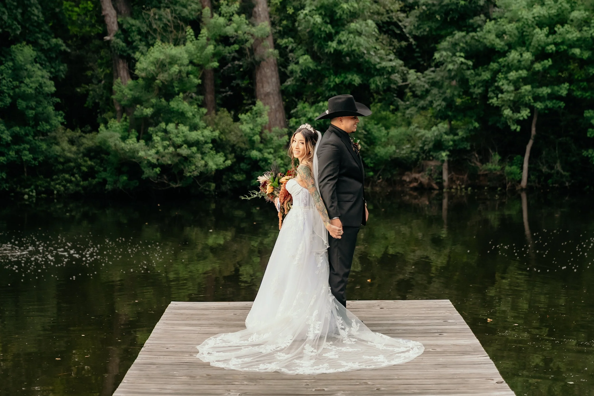 A bride and groom holding hands on a wooden dock by a river, surrounded by lush green trees, during their wedding.