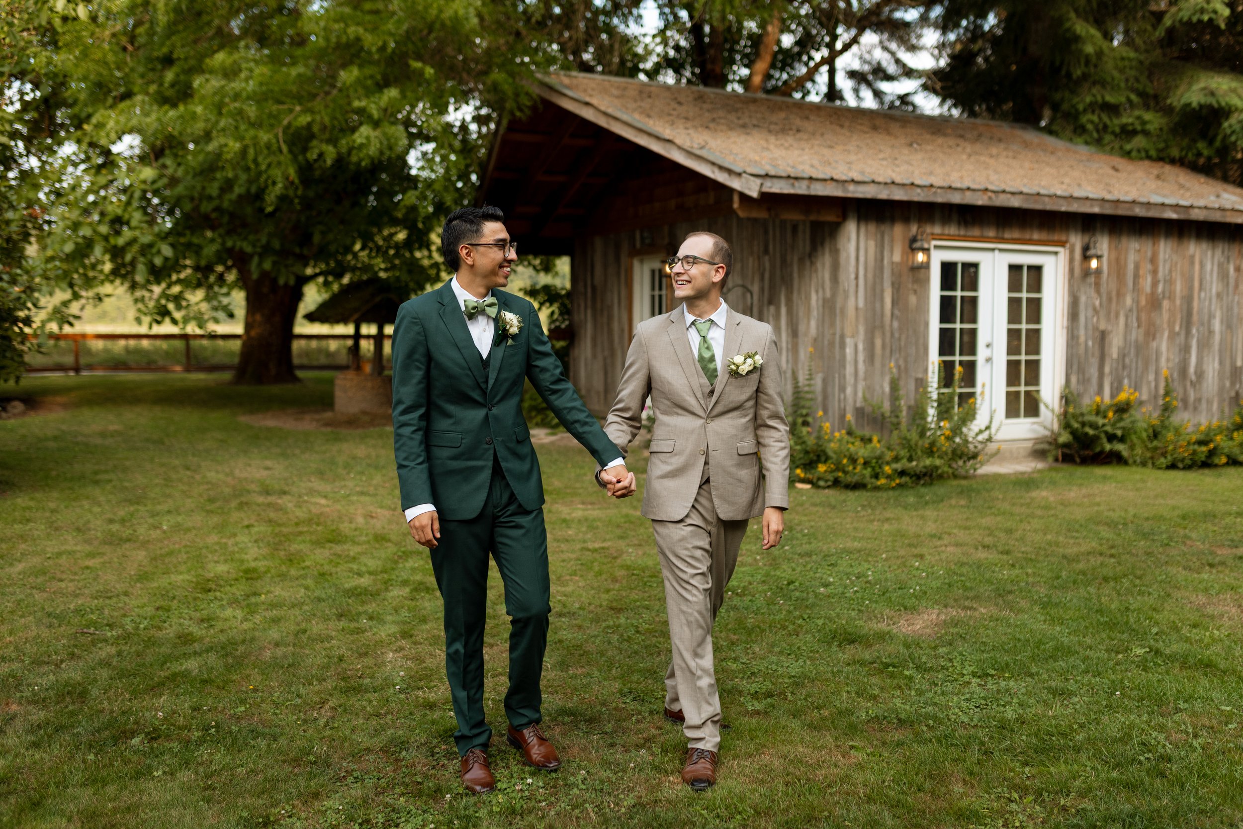 Two men in suits holding hands and smiling while walking on grass outside with a wooden house and trees in the background.