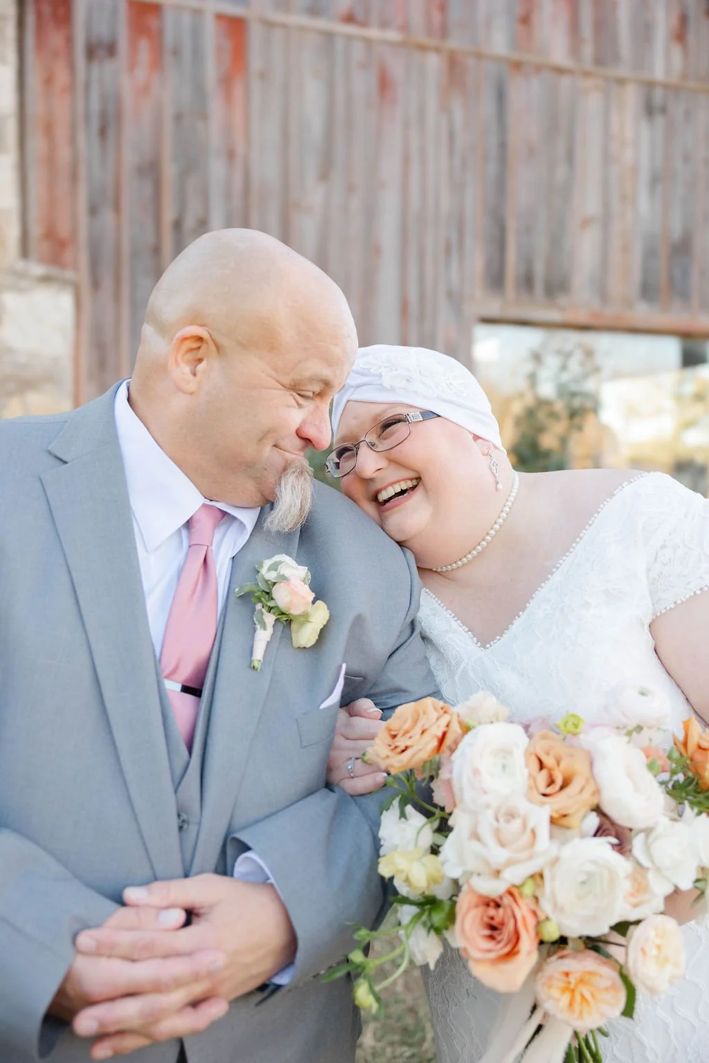 A couple sharing a joyful moment at their wedding, with the man dressed in a light gray suit and pink tie, and the woman in a white dress and headscarf, holding a bouquet of light pink, peach, and white flowers.