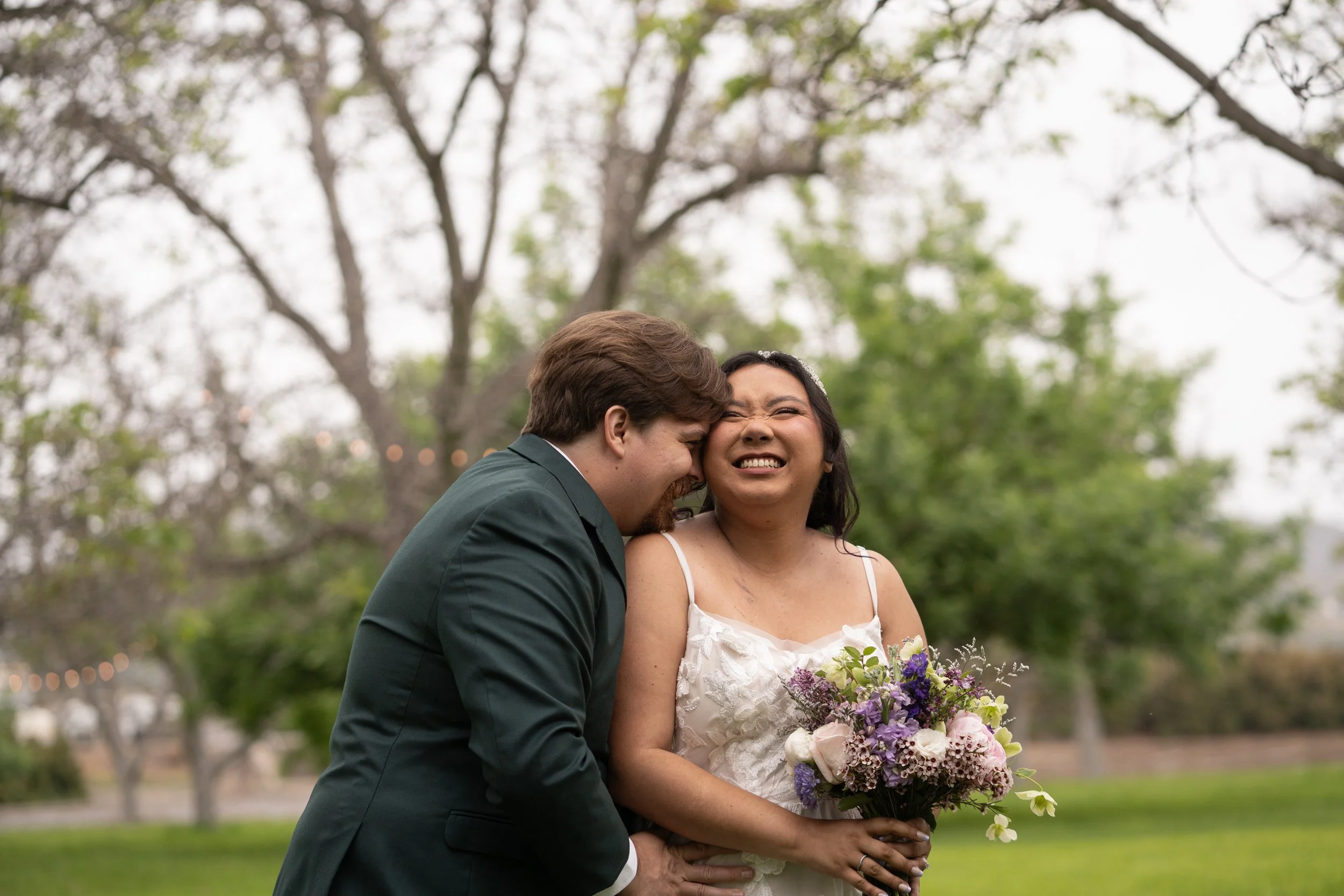 A newlywed couple stands outdoors under a tree, sharing a joyful moment. The woman is holding a bouquet of flowers and smiling, while the man leans in with a happy expression.