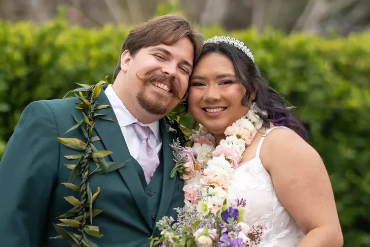 A newlywed couple is smiling and posing outdoors, wearing wedding attire and floral leis.