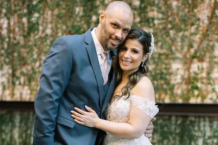 A newlywed couple, a man in a blue suit and a woman in a white lace wedding dress, smiling and embracing outdoors with trees in the background.