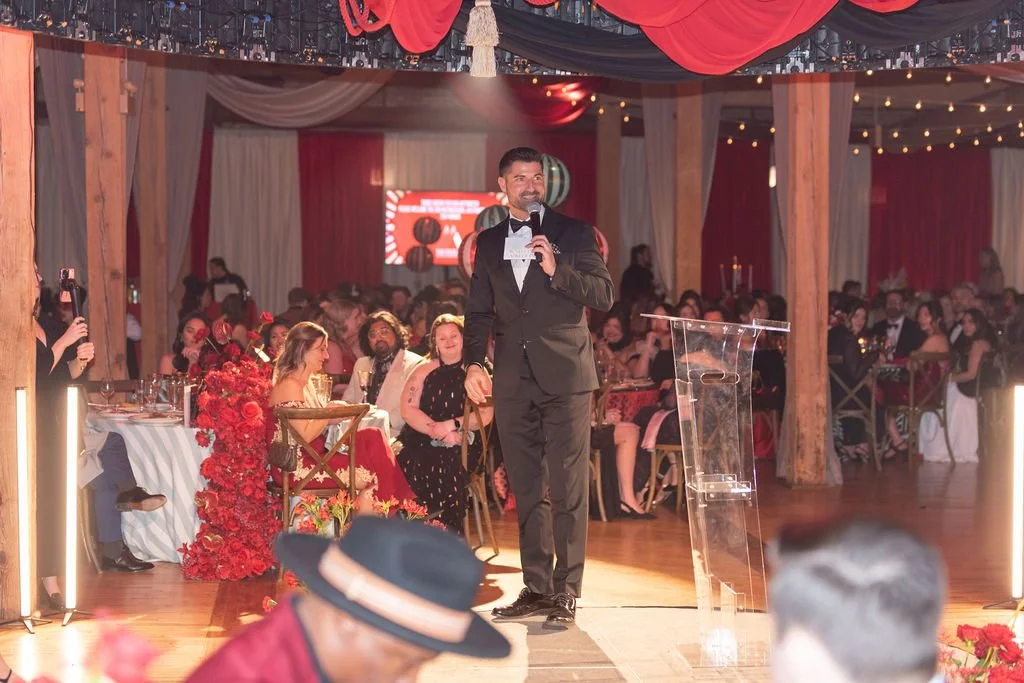 Man in a black tuxedo giving a speech at a gala reception with guests seated at decorated tables in the background.