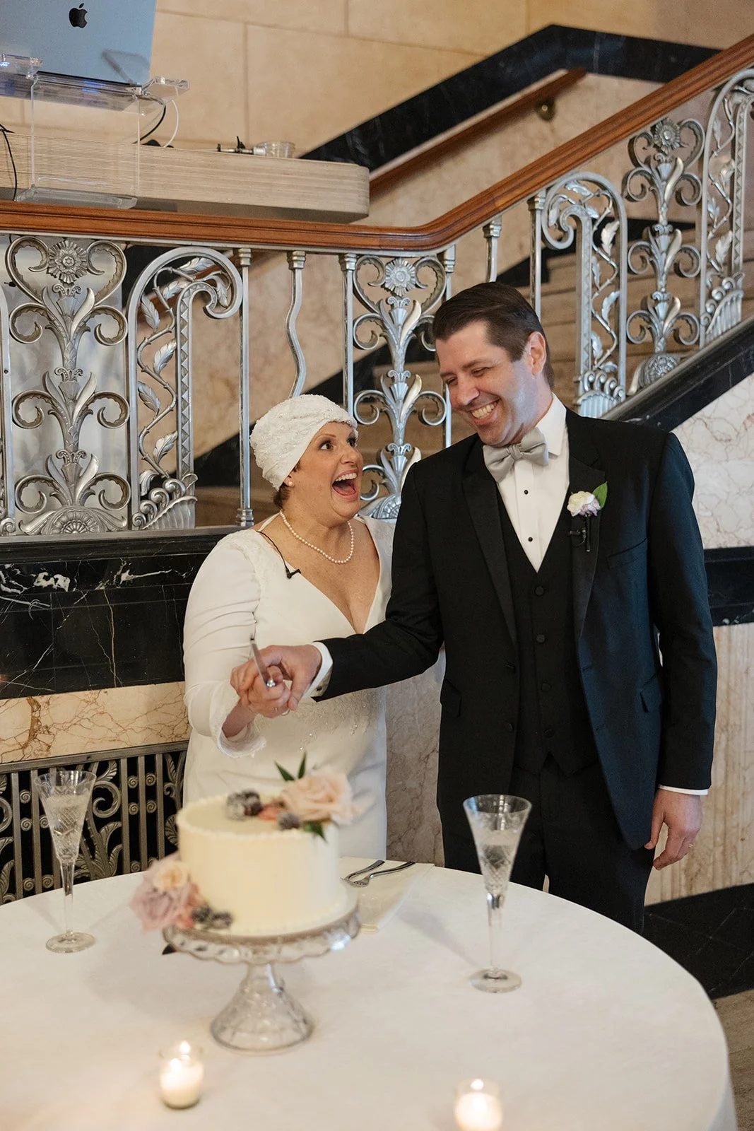 A woman and a man at a wedding reception, smiling and holding hands over a table with a wedding cake, in an elegant decorated venue.