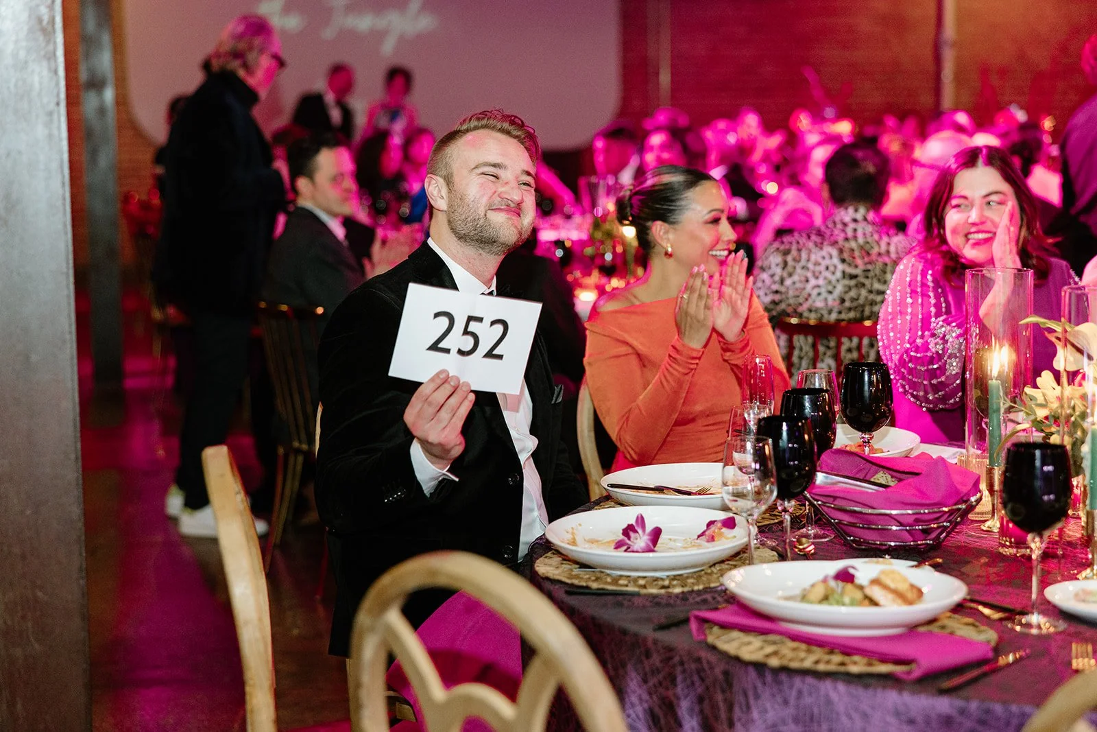 People sitting at a banquet table during an event, one man holding a sign with the number 252, surrounded by food and glasses, with pink lighting and decorations in the background.