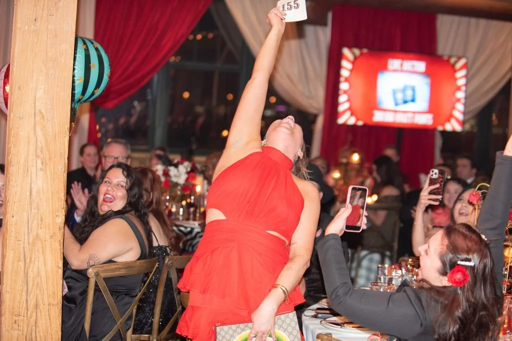Woman in a red dress winning an award or game at a festive event, holding a white card above her head while others cheer and take photos.