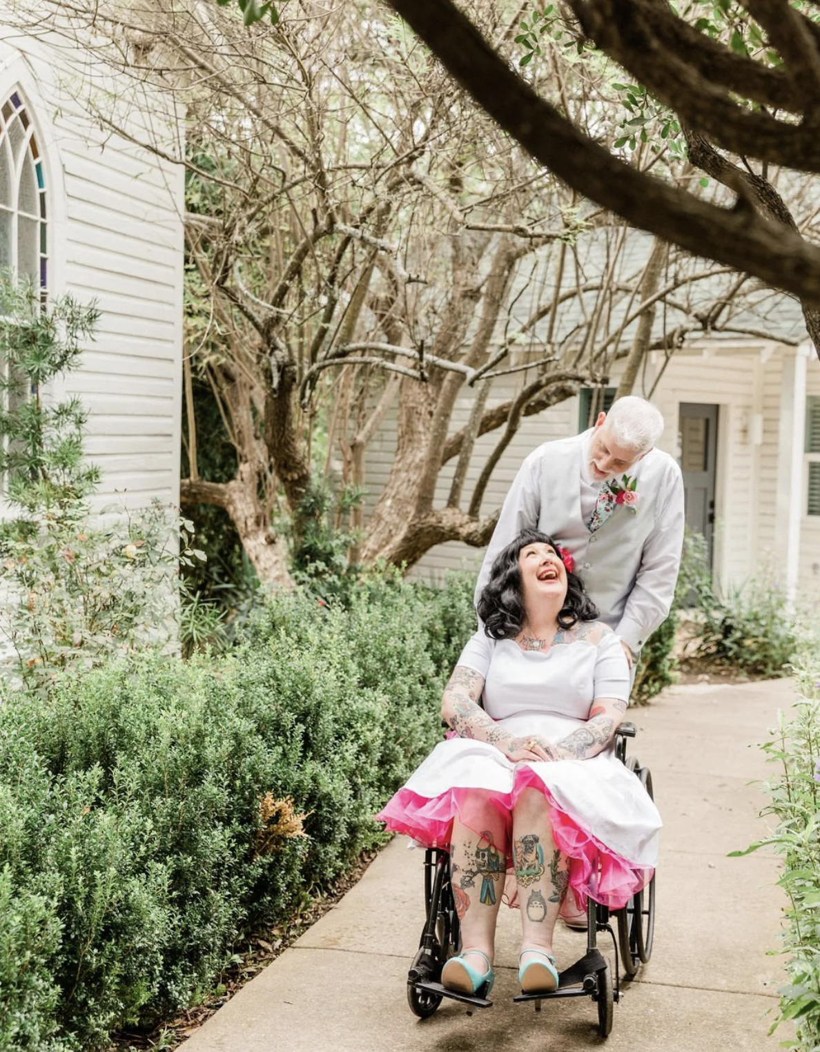A woman in a wheelchair with tattoos and dark hair, wearing a white dress with pink accents, smiling at a man with gray hair in a light gray vest, who is leaning toward her and smiling. They are outdoors on a garden path surrounded by greenery and trees, with a house in the background.