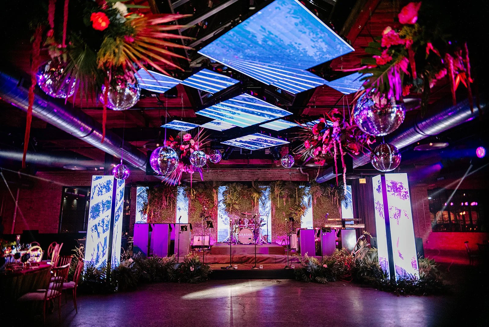 An event space decorated with pink flowers and hanging disco balls, featuring a stage with musical instruments and vertical screens displaying a blue forest silhouette.