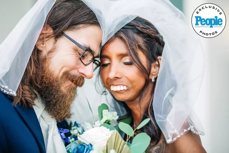 A bride and groom sharing a close moment under a veil at their wedding, with the bride smiling and the groom winking, holding a bouquet of flowers.
