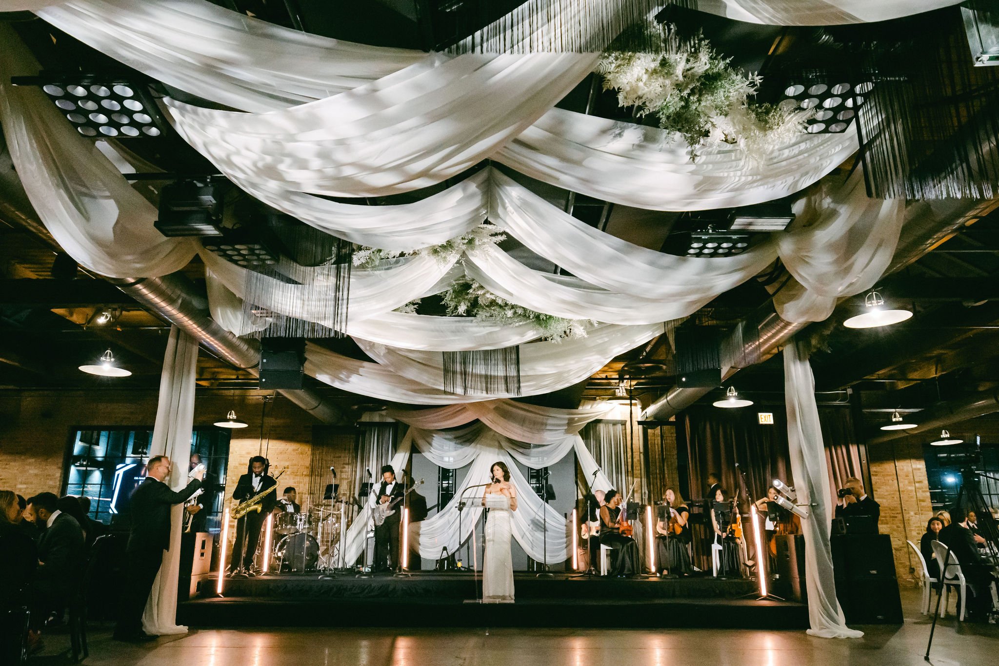 An indoor wedding reception with a live jazz band performing on a decorated stage with white drapes and floral arrangements, guests seated at tables, and some standing near the band.