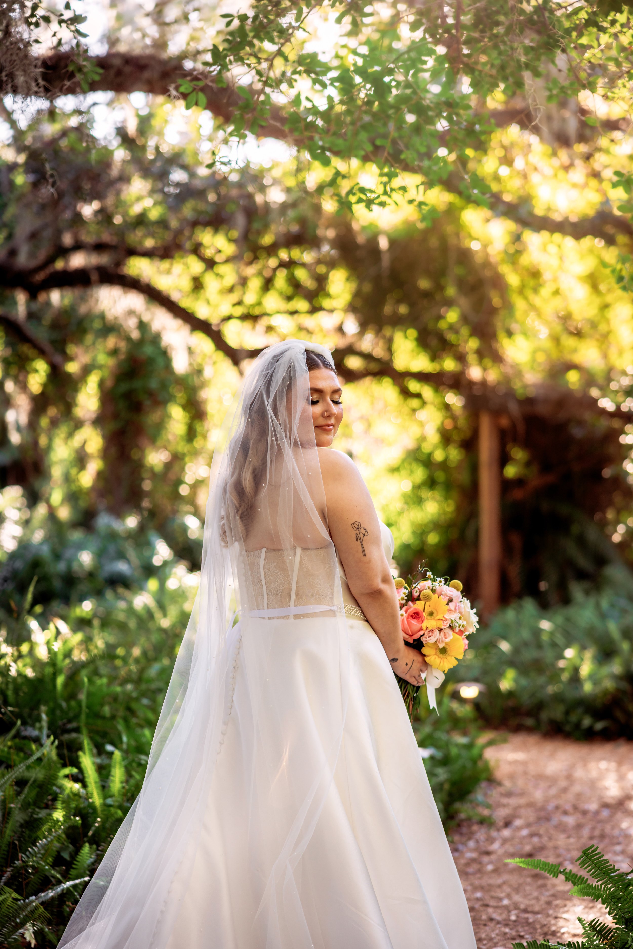 A bride in a white wedding dress and veil holds a bouquet of pink, yellow, and peach flowers in an outdoor garden setting, with sunlight filtering through the trees.