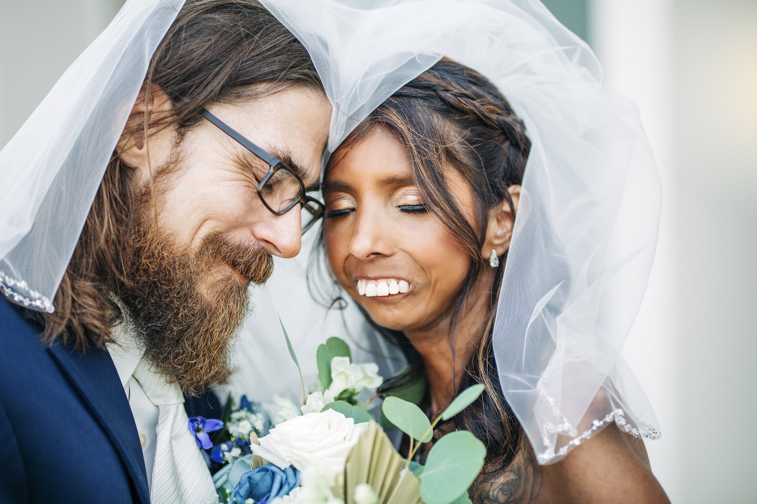 A man with glasses and a beard and a woman with dark hair and earrings, both dressed up, kissing under a white veil during a wedding ceremony, with the woman holding a bouquet of white roses and green foliage.
